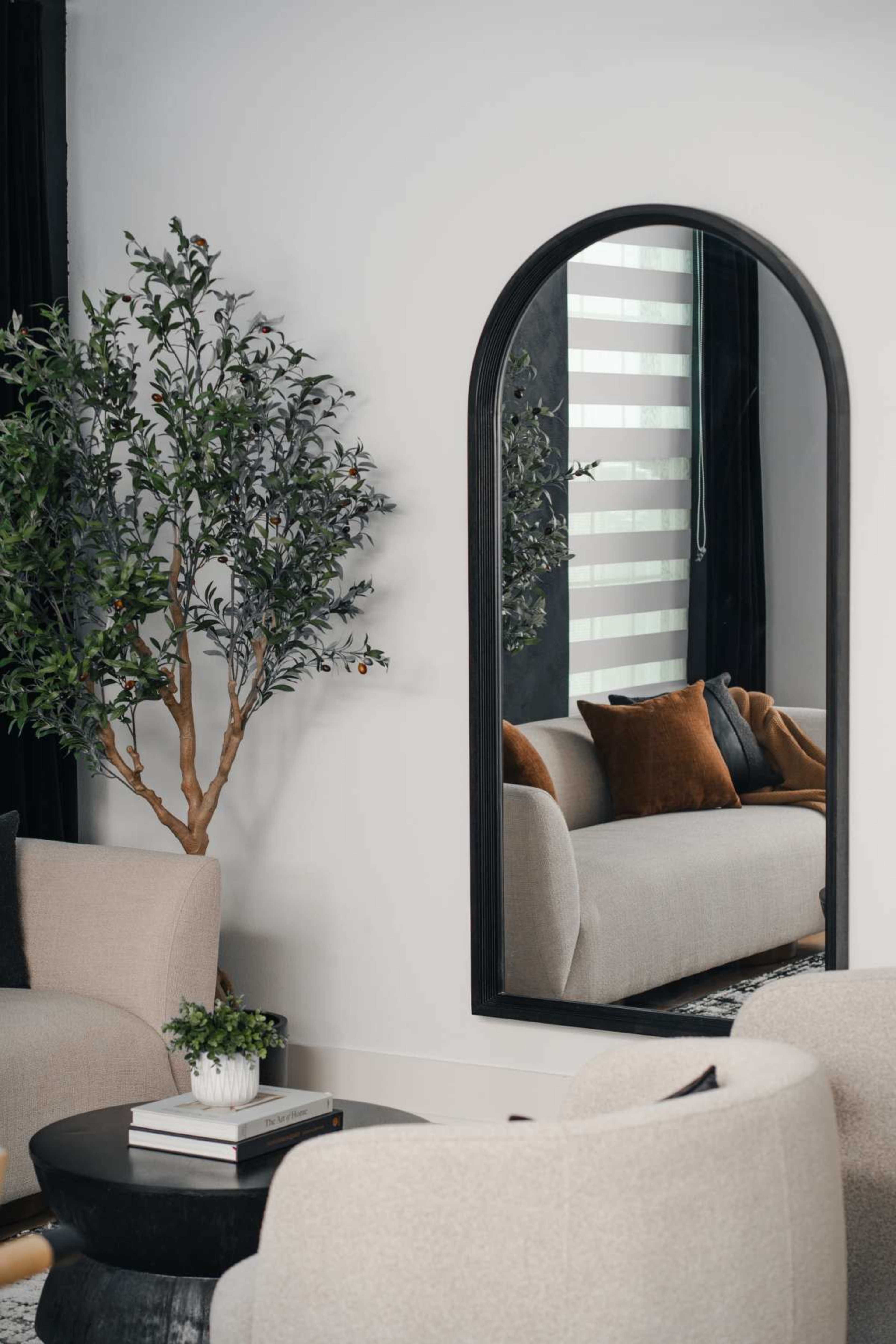 A cozy living room features two beige armchairs, a black side table with books, and a large mirror reflecting a couch and a decorative plant.