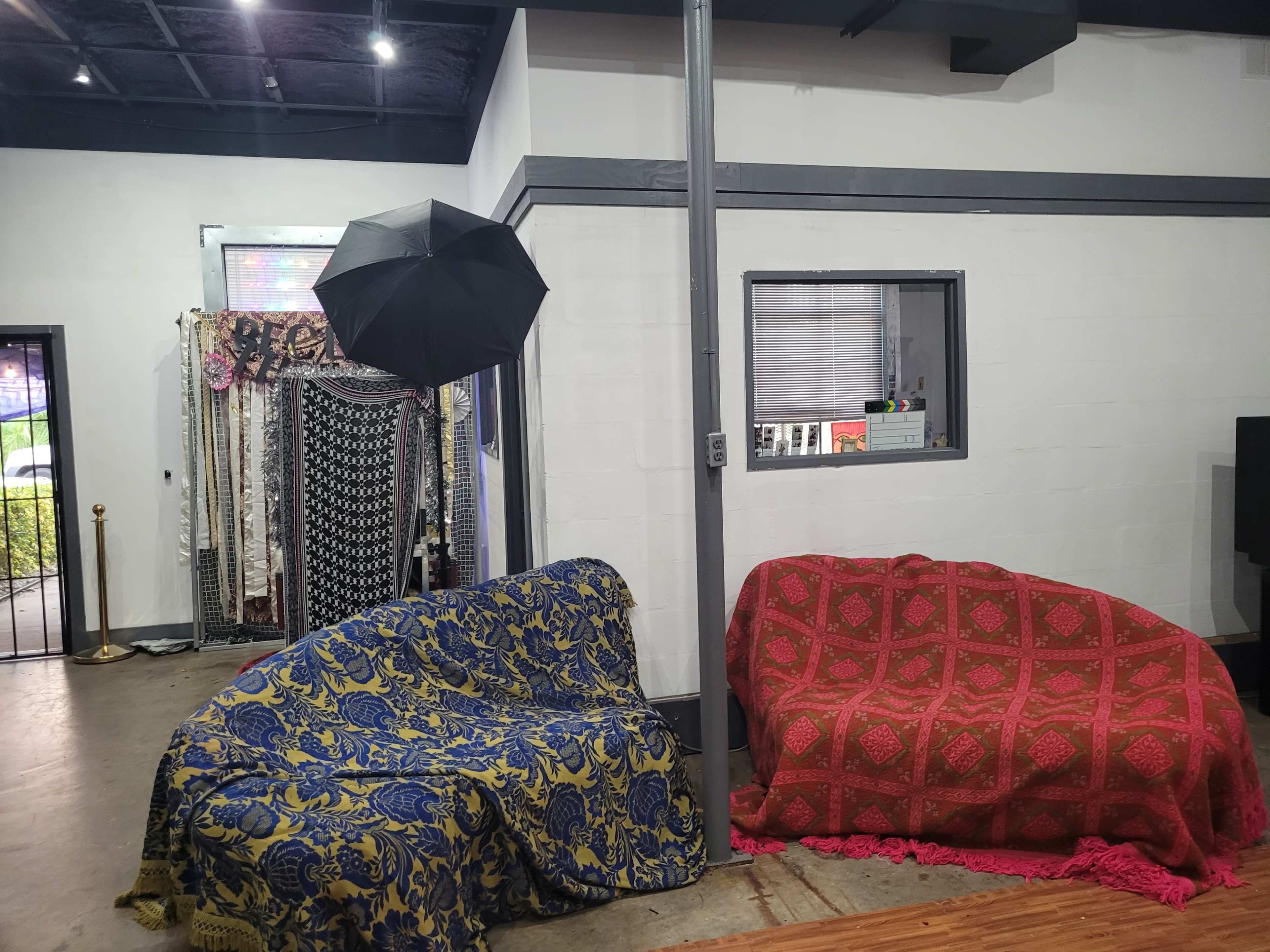 The image shows two covered sofas, one blue and gold and the other red, positioned in a room with a black umbrella standing nearby and patterned fabrics hanging on the wall.