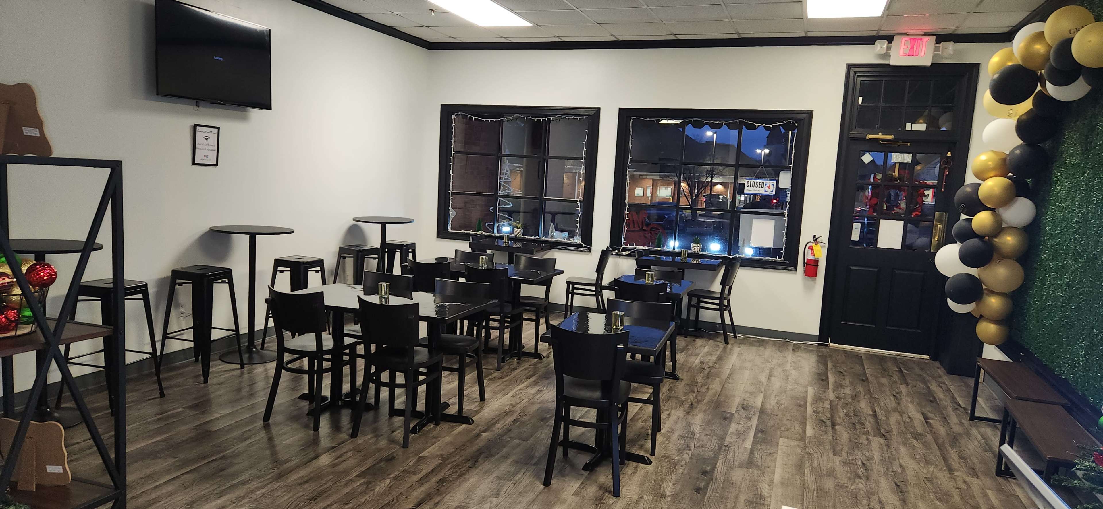 The image shows a modern, sparsely furnished café with black tables and chairs arranged near two windows, and a decorative wall with balloons.