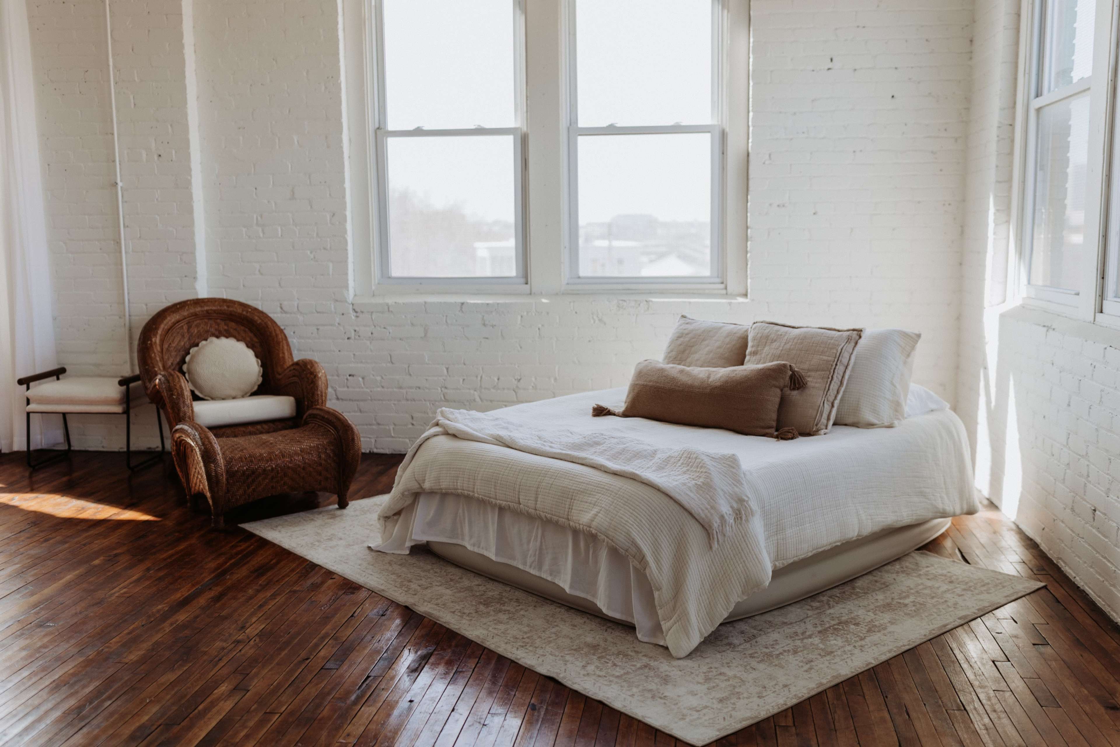 The image features a bright bedroom with a wooden floor, a large window letting in natural light, a neatly made bed with decorative pillows, and a woven chair in the corner.