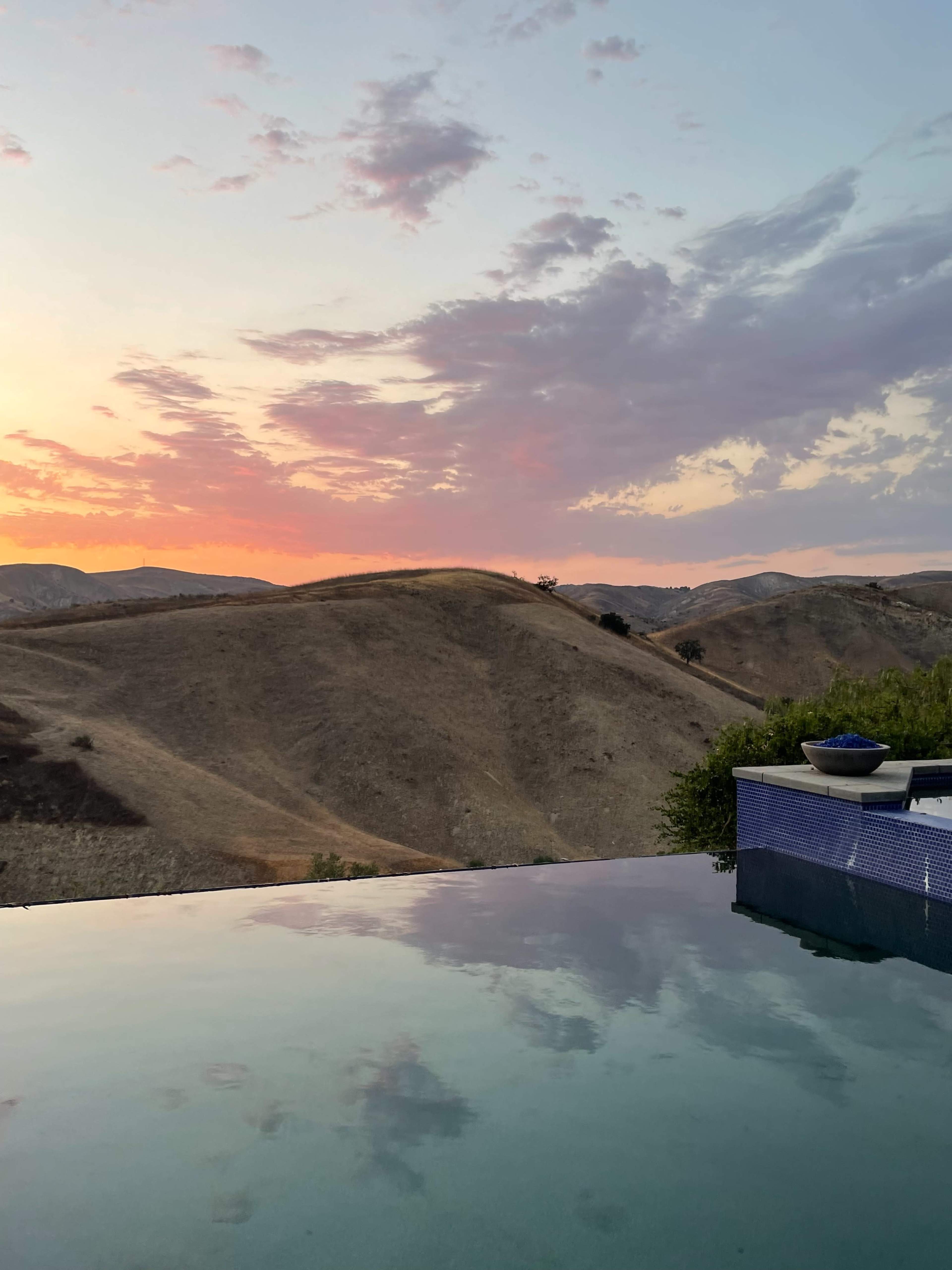 The image shows a tranquil pool reflecting a colorful sunset over dry, hilly terrain.