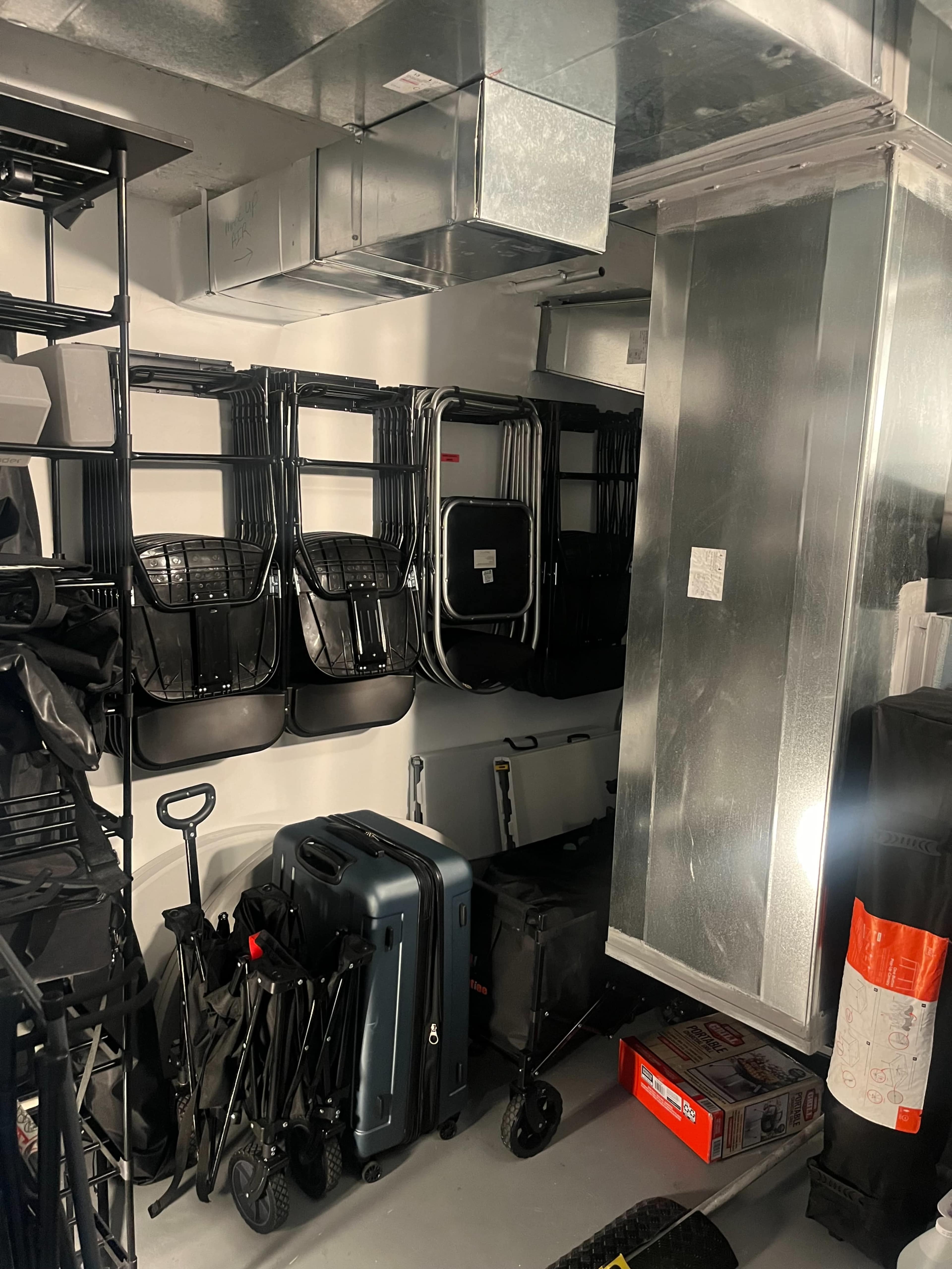 A storage area is filled with metal shelving, stacked folding chairs, various pieces of luggage, and ventilation ducts.