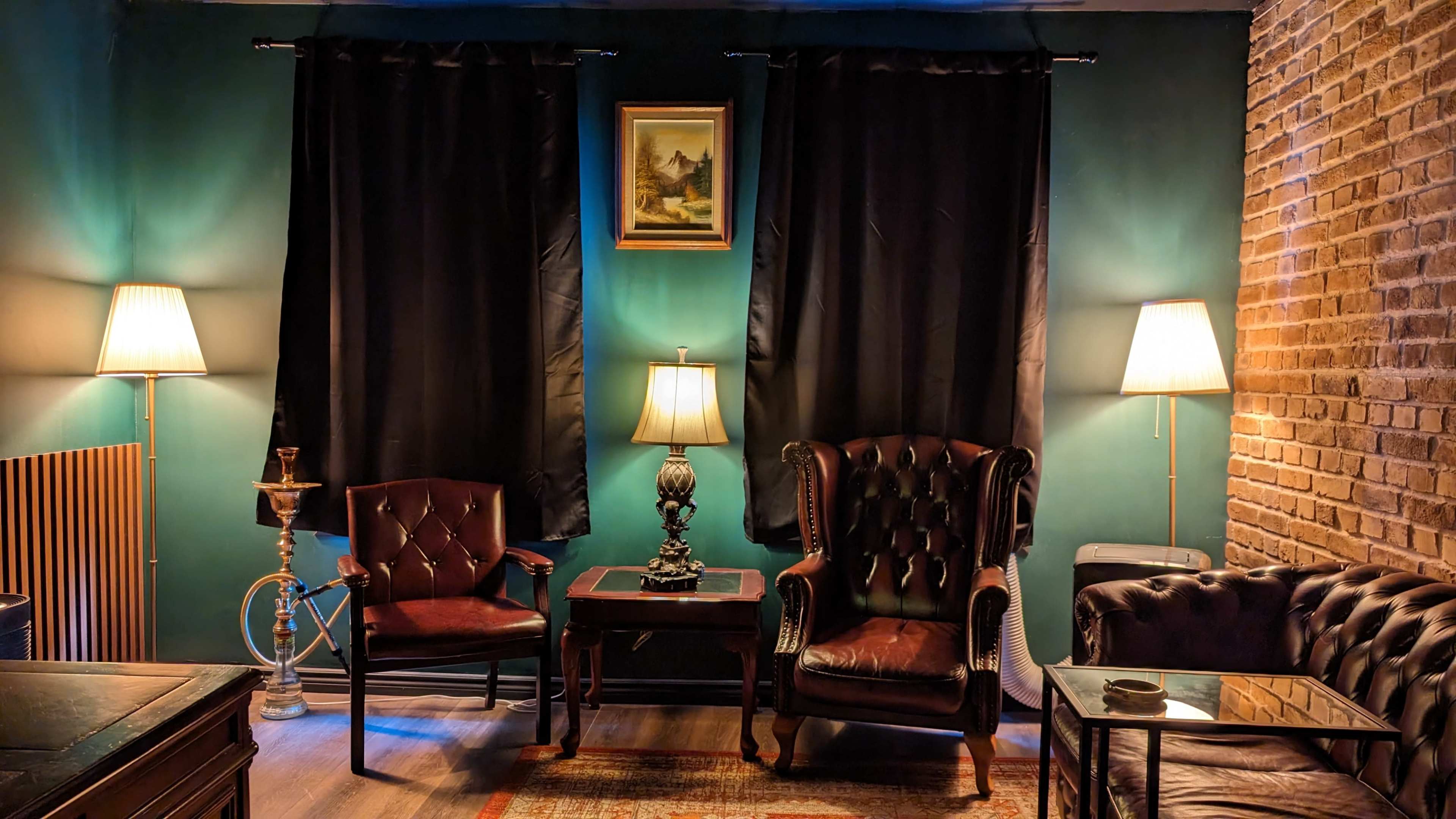 A cozy sitting area features two leather armchairs, a small wooden table, and two floor lamps beside dark curtains and a brick wall.