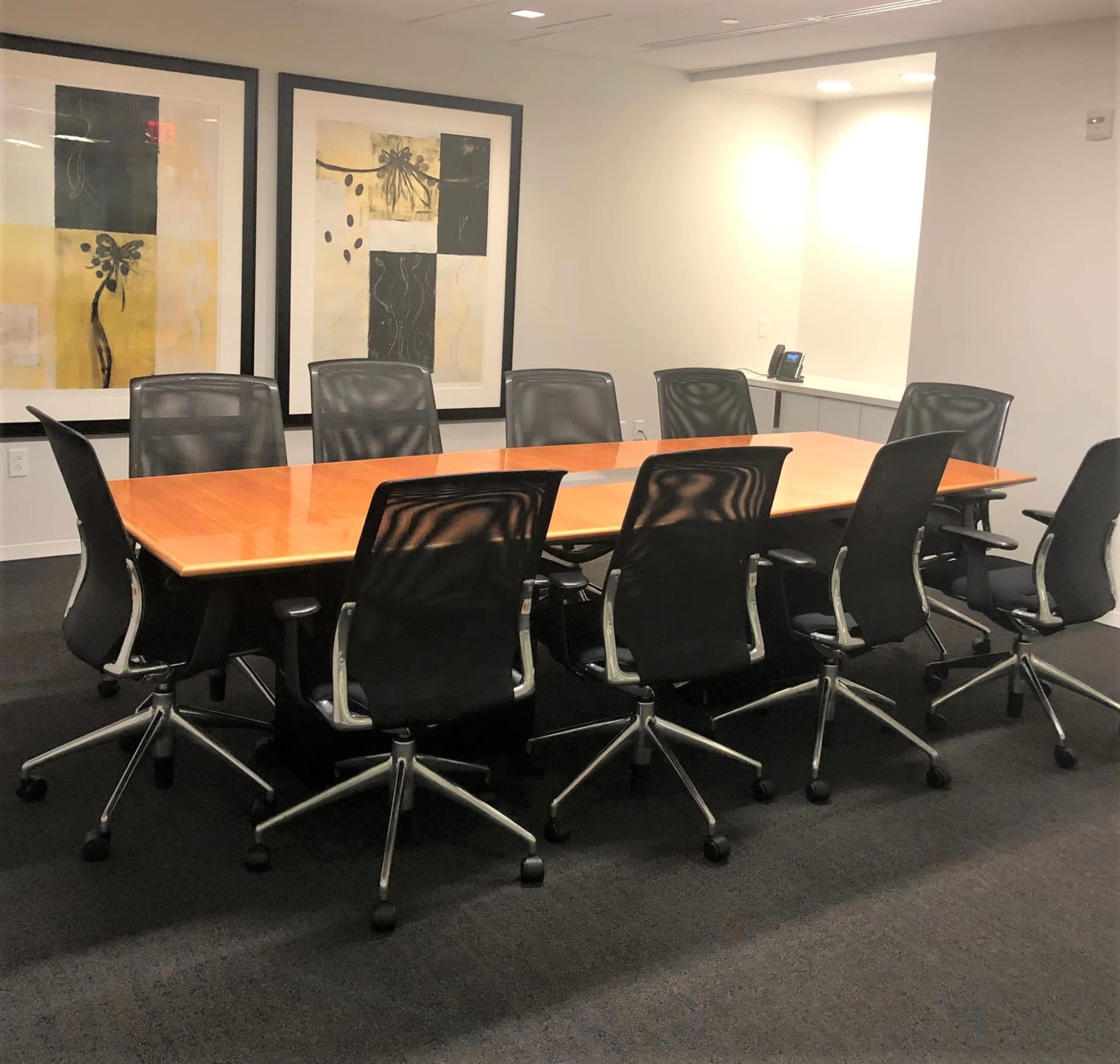 A long conference table surrounded by ergonomic chairs is situated in a well-lit office meeting room, complemented by two framed artworks on the walls.