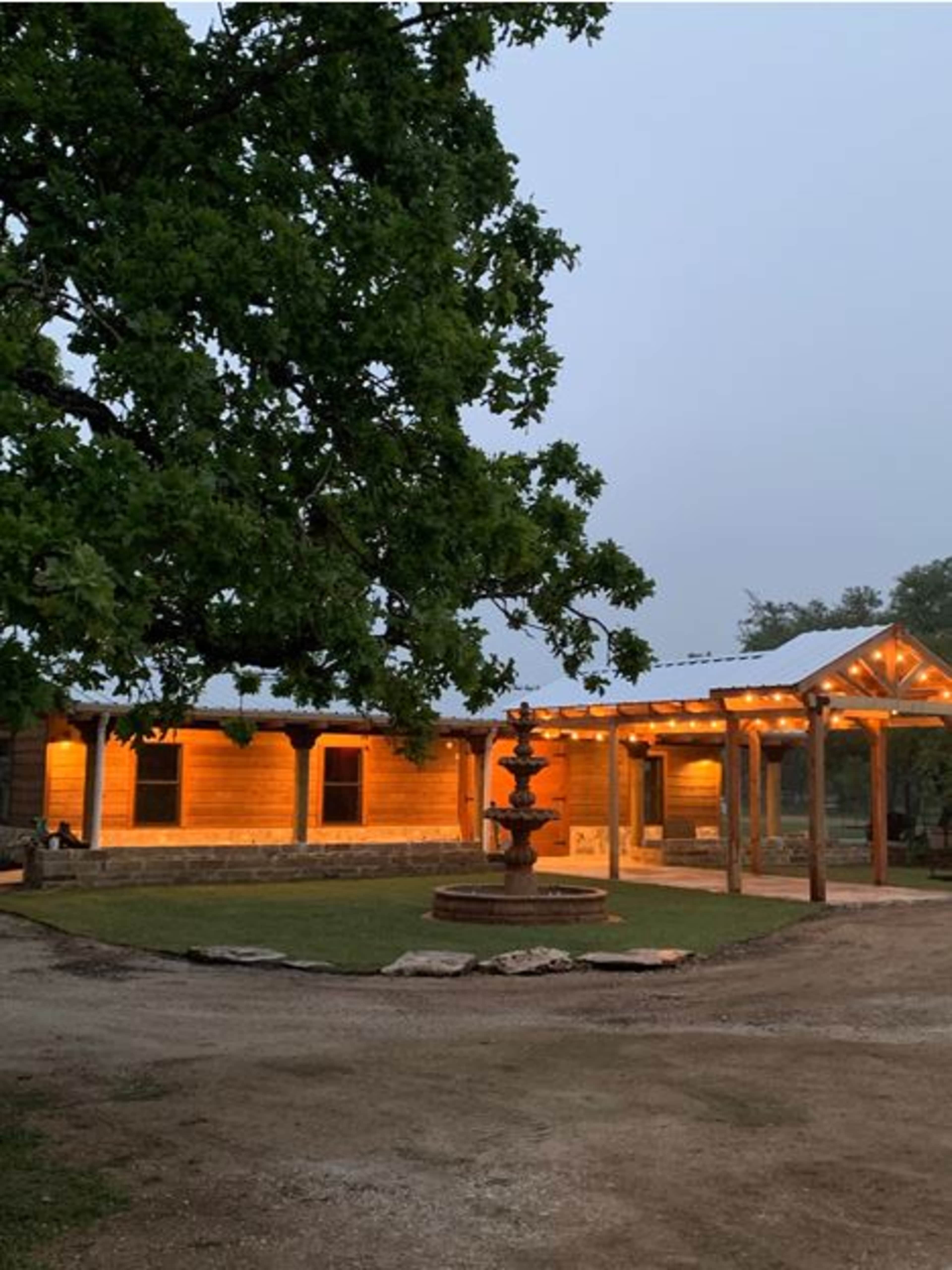 A wooden ranch-style house with a stone accent, a fountain, and covered porch is surrounded by grass and trees in a misty evening setting.