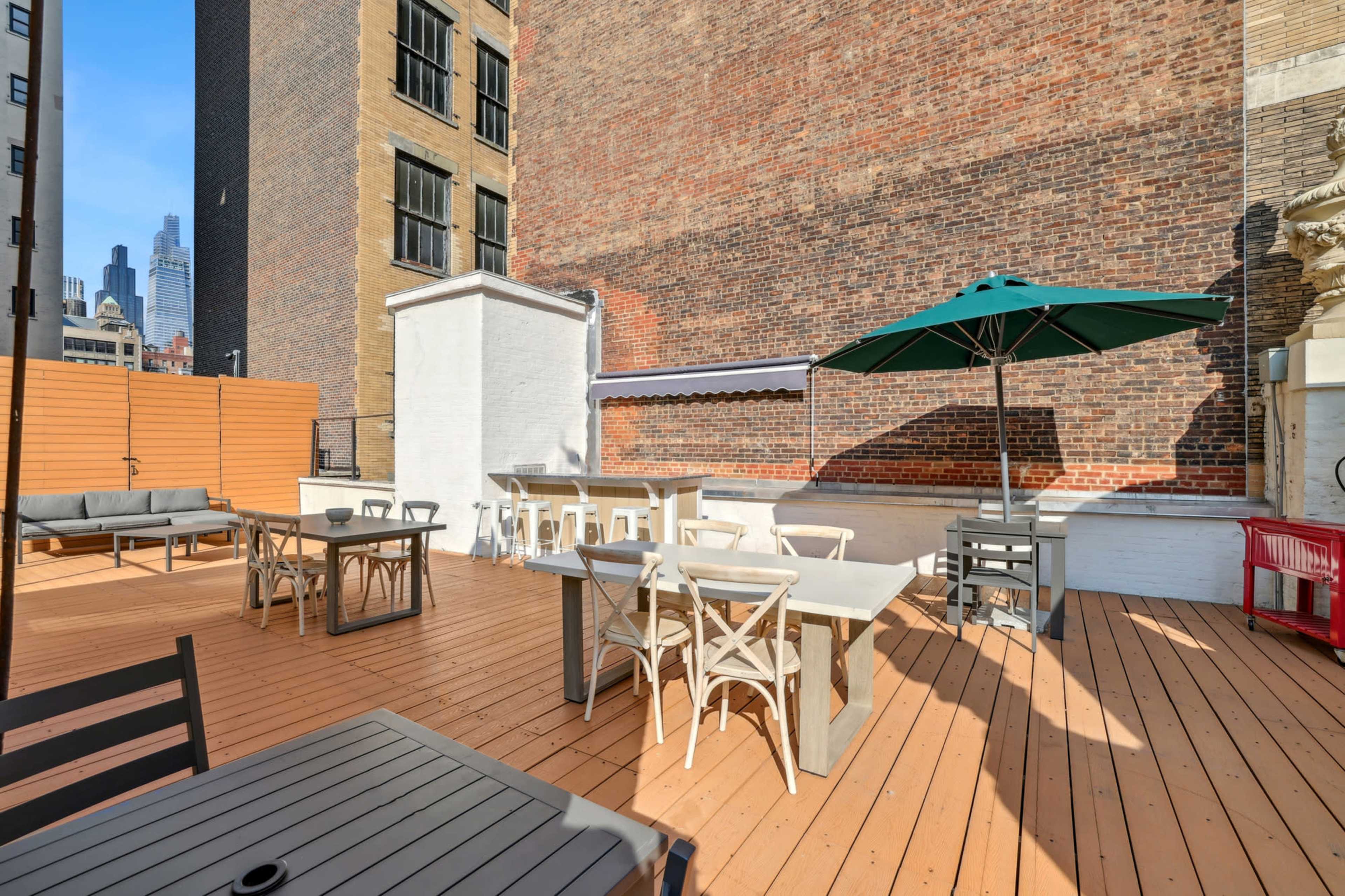The image shows a rooftop terrace area featuring wooden decking, several tables and chairs, and an umbrella.