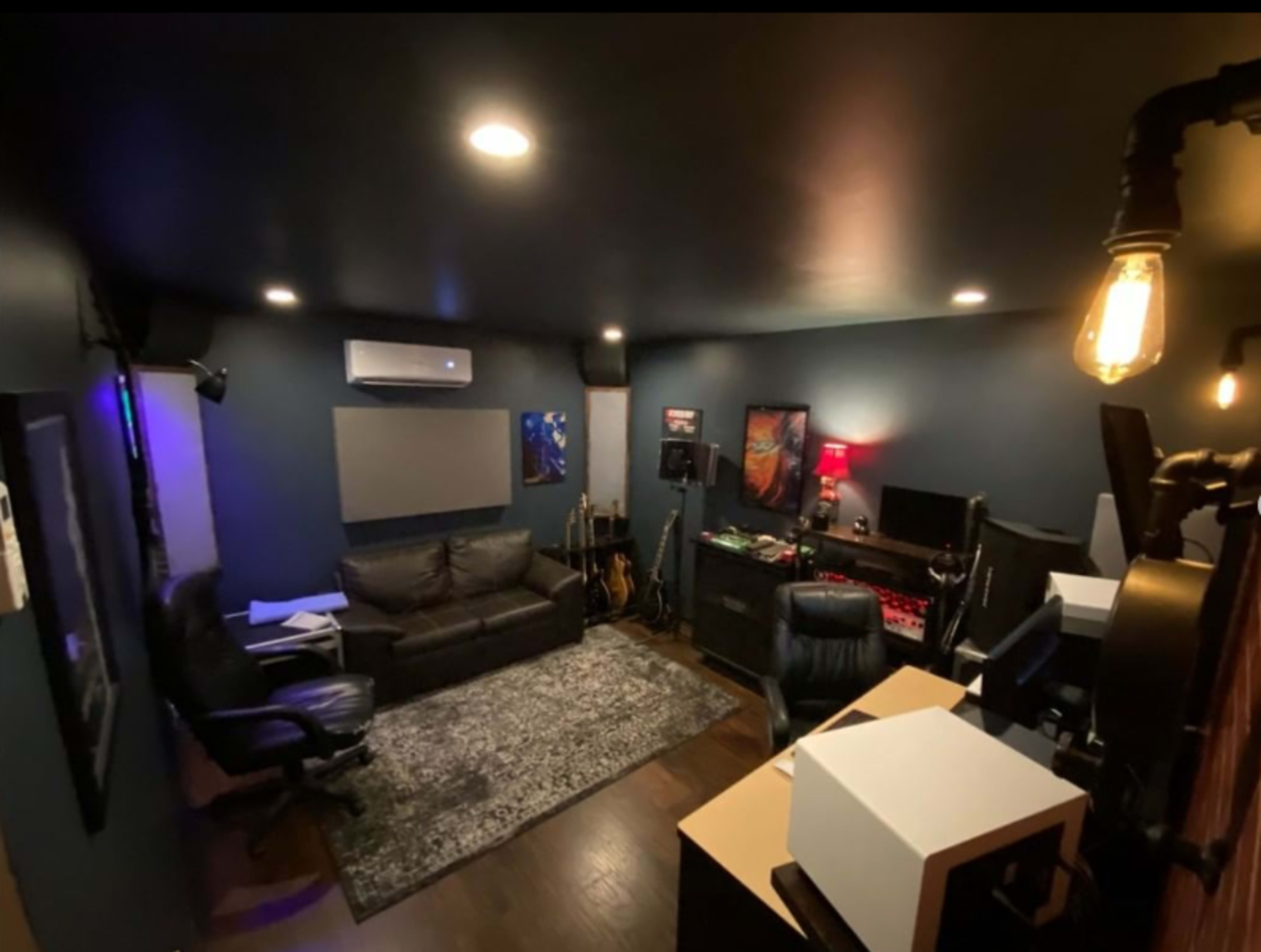 A small recording studio with dark walls, a sofa, various musical instruments, and studio equipment arranged around the space.