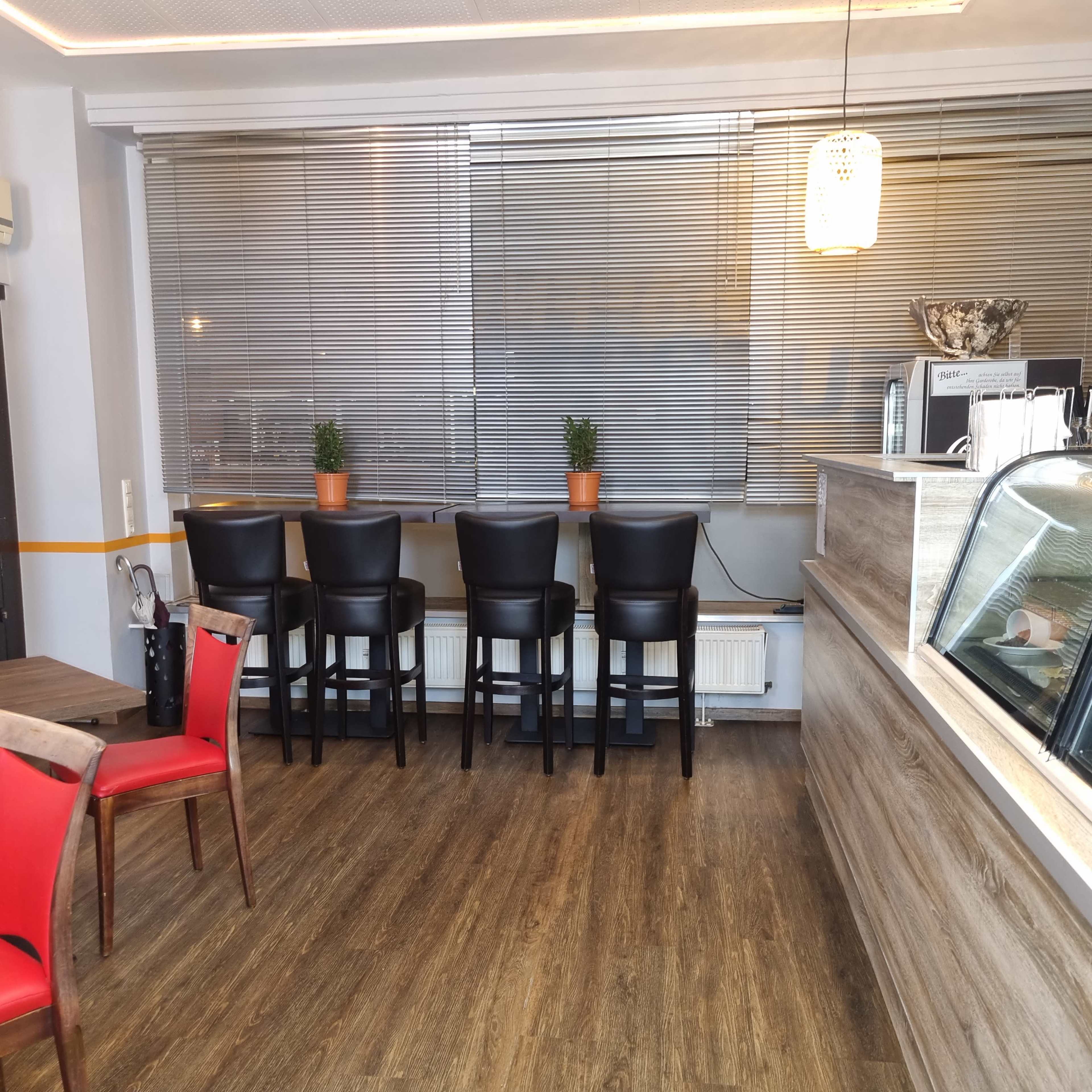 The image shows a modern café interior featuring a counter on the right, tall black chairs lined up against a window, and a few small potted plants on the ledge.