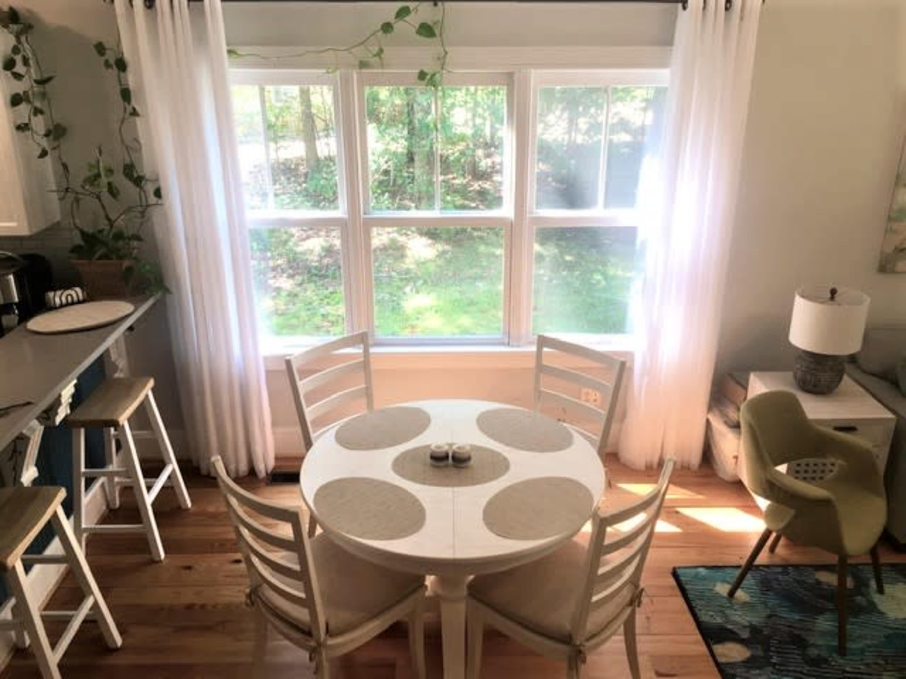 A round dining table with four chairs is positioned near large windows that overlook a green outdoor area, with light streaming in through sheer curtains.
