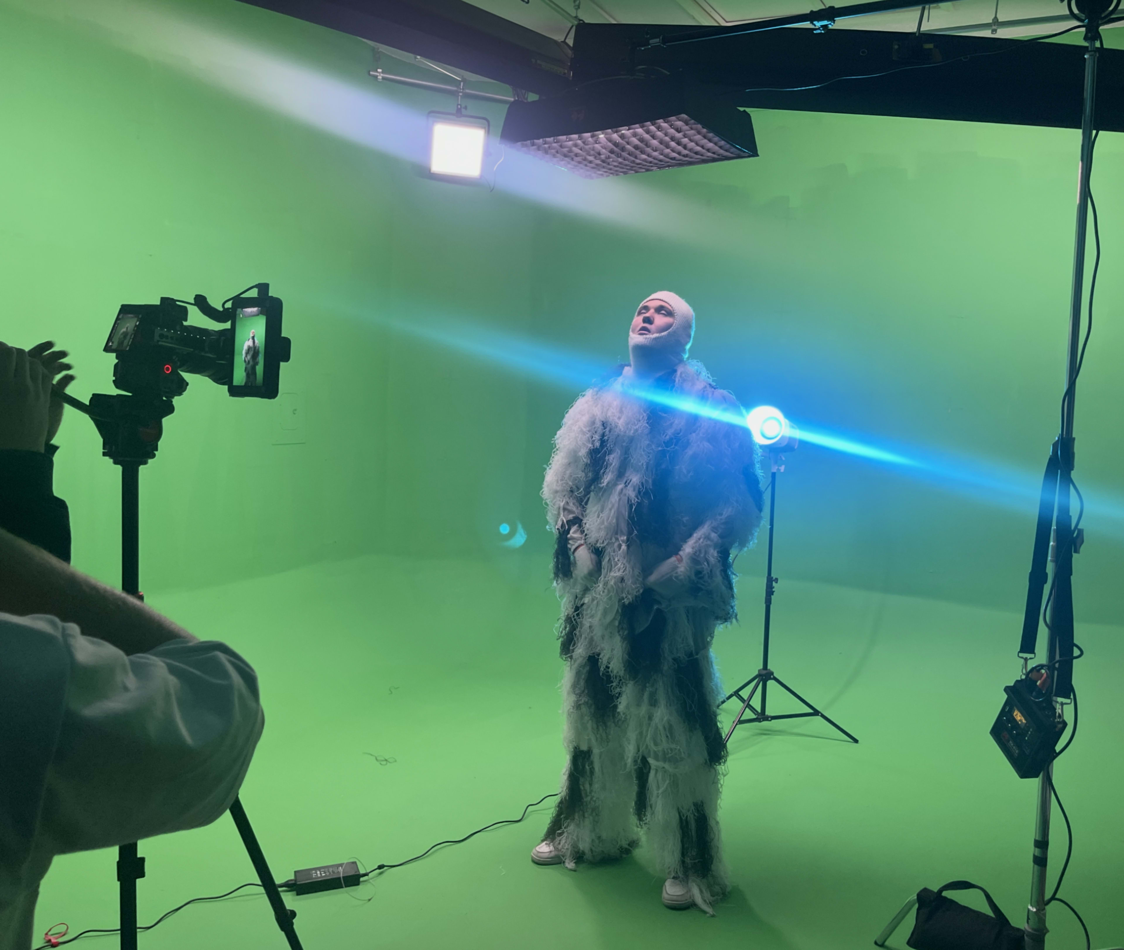 A person in a feathery outfit stands in a green studio while being photographed with bright lights directed at them.
