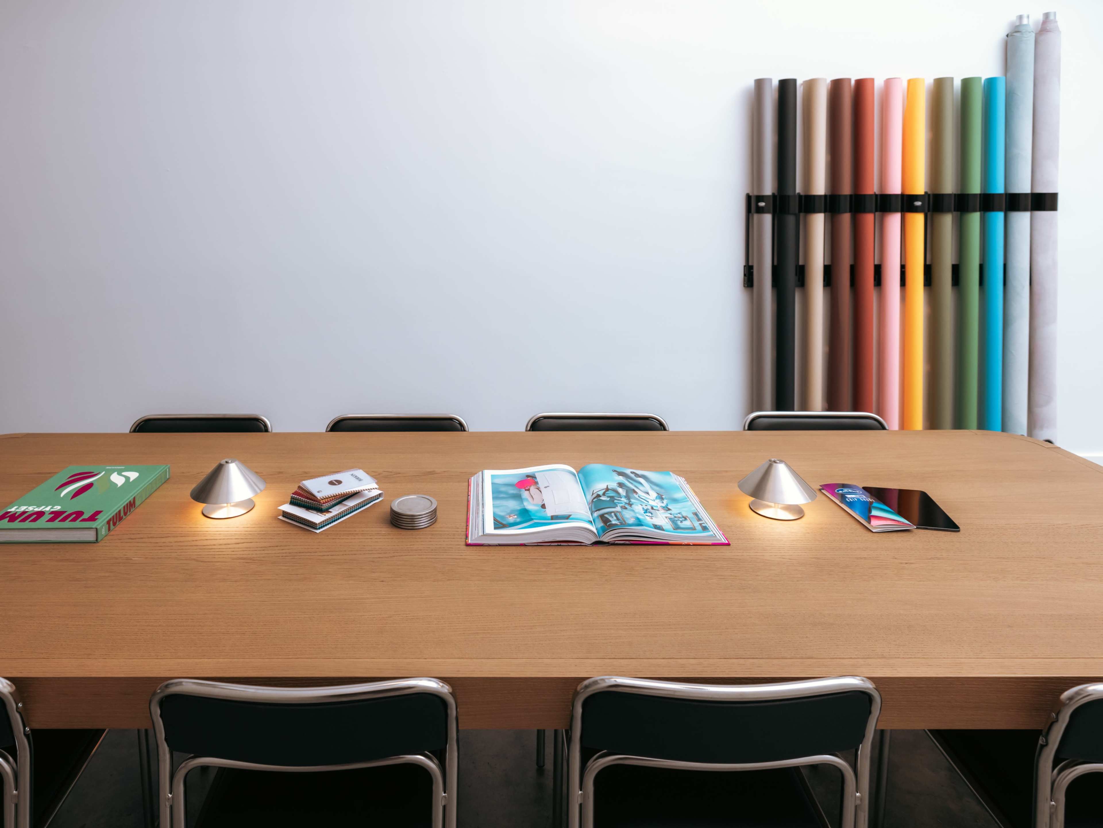 A wooden table with several chairs features open magazines, a tablet, and two small lamps, while colorful rolls of paper are neatly arranged on the wall behind.