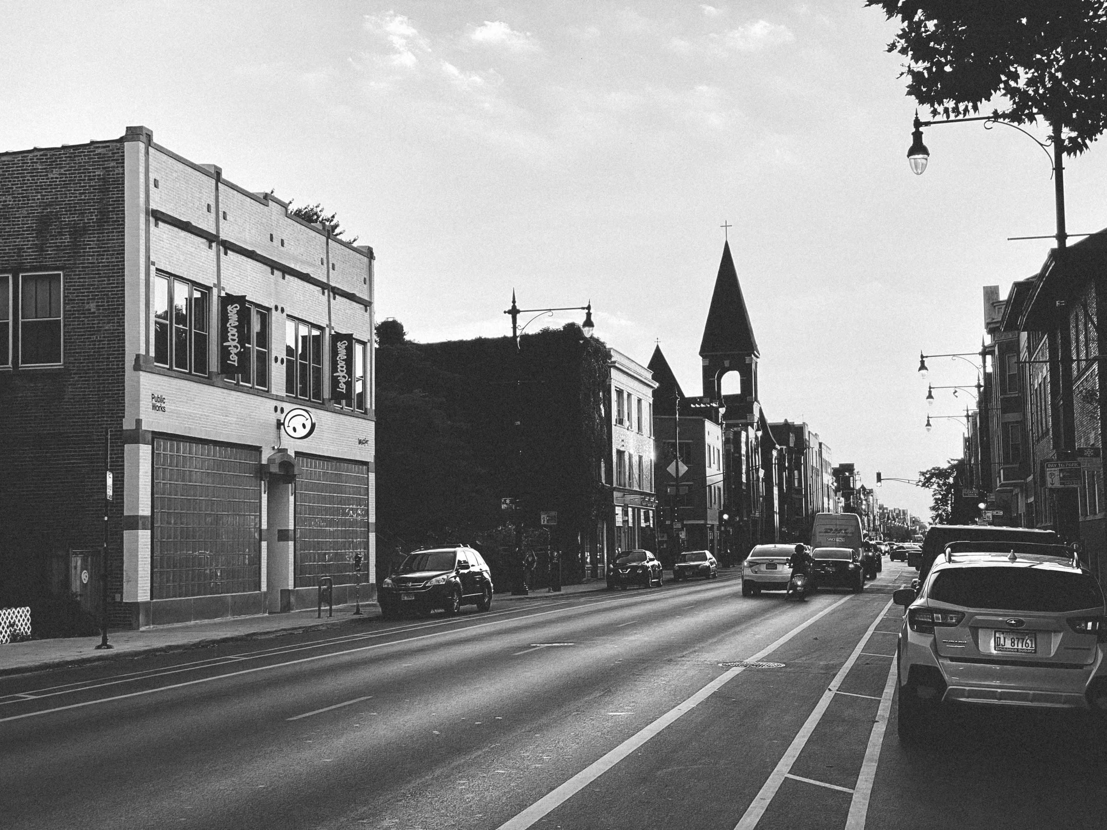 A wide street with vehicles parked along the sides, featuring a mix of buildings, including a church with a tall spire in the background.