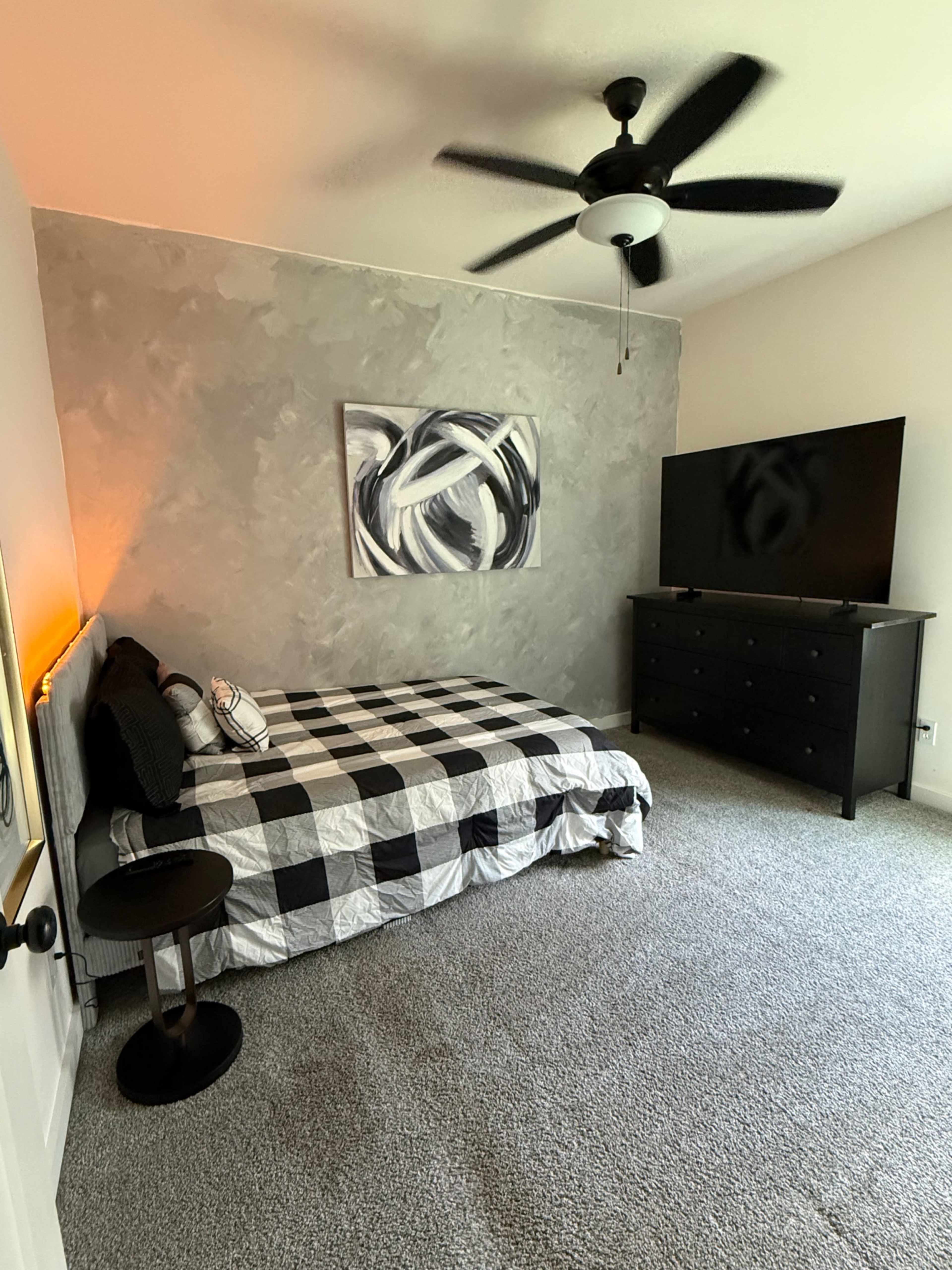 The image shows a bedroom with a checkered bedspread, a black dresser, a large wall-mounted TV, and a ceiling fan.
