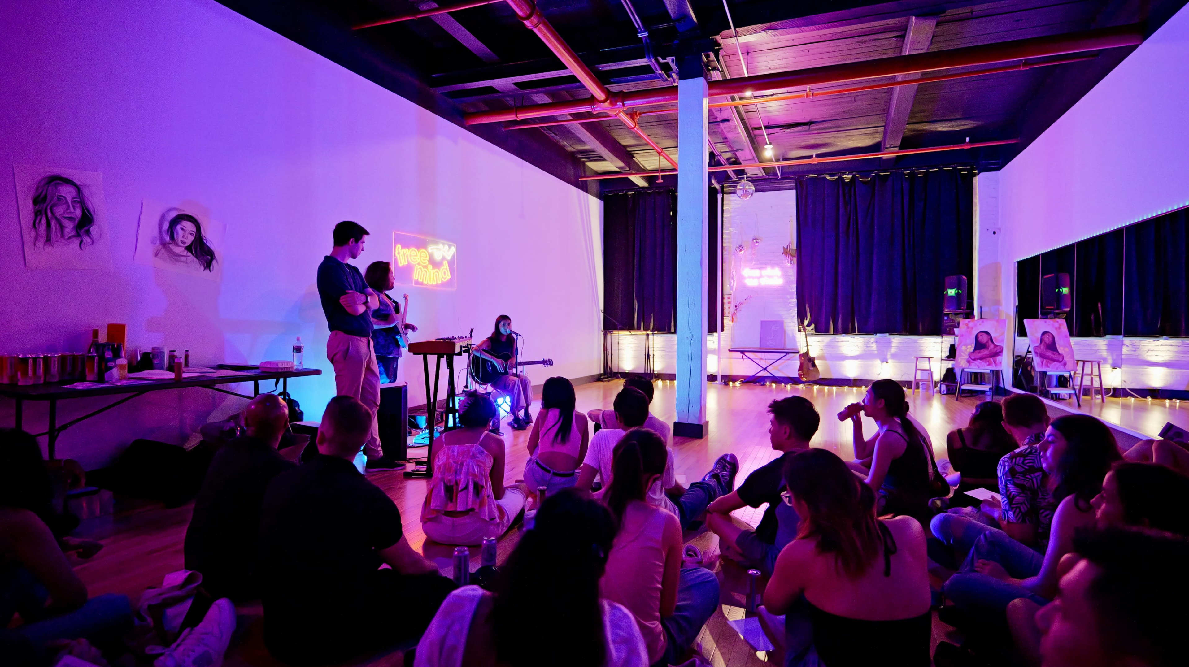 A group of people is seated on the floor in a dimly lit space, watching a musician perform with a guitar on a small stage.