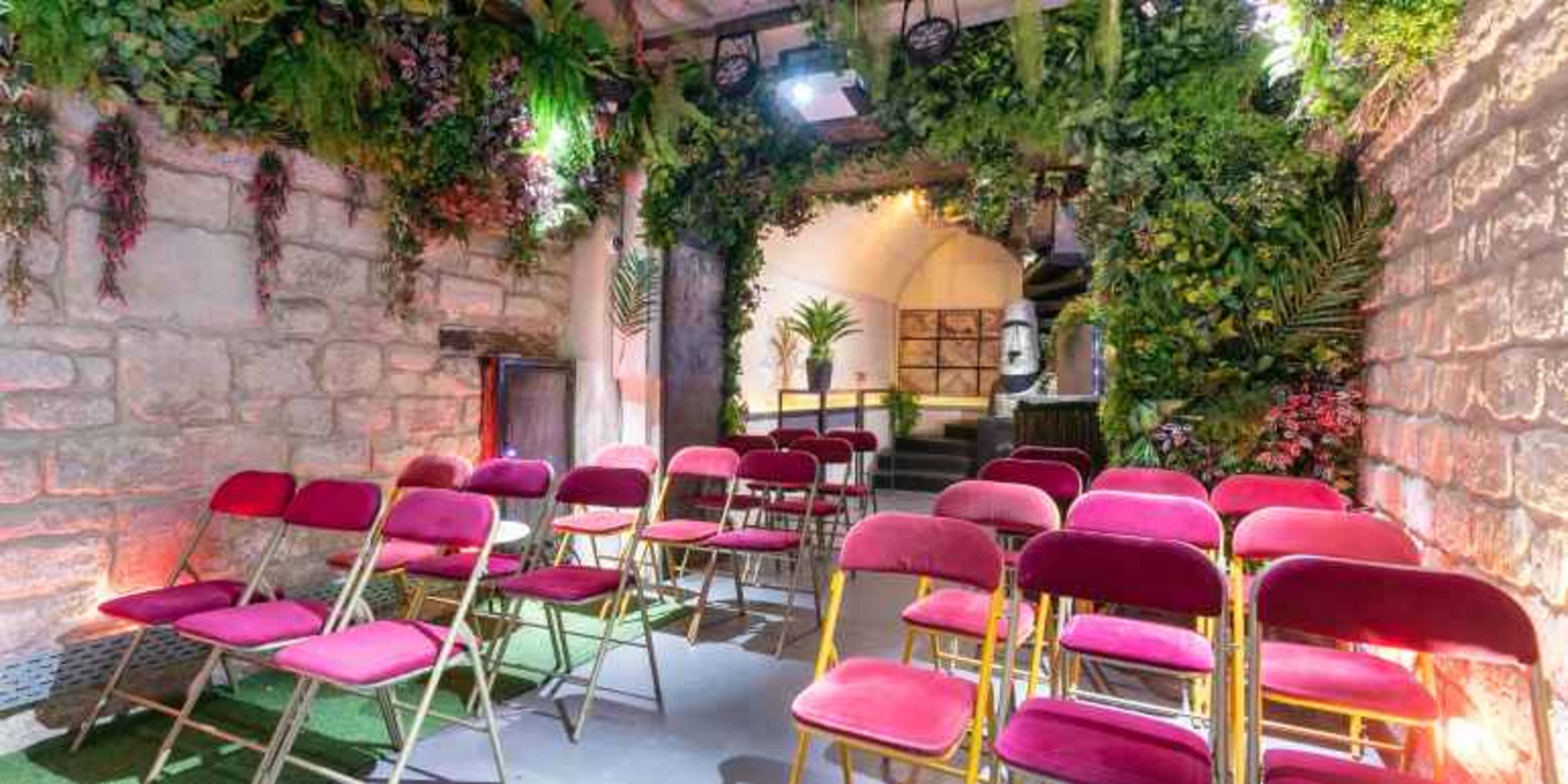 A small indoor space features rows of pink chairs set up in front of a backdrop adorned with greenery and stone walls.