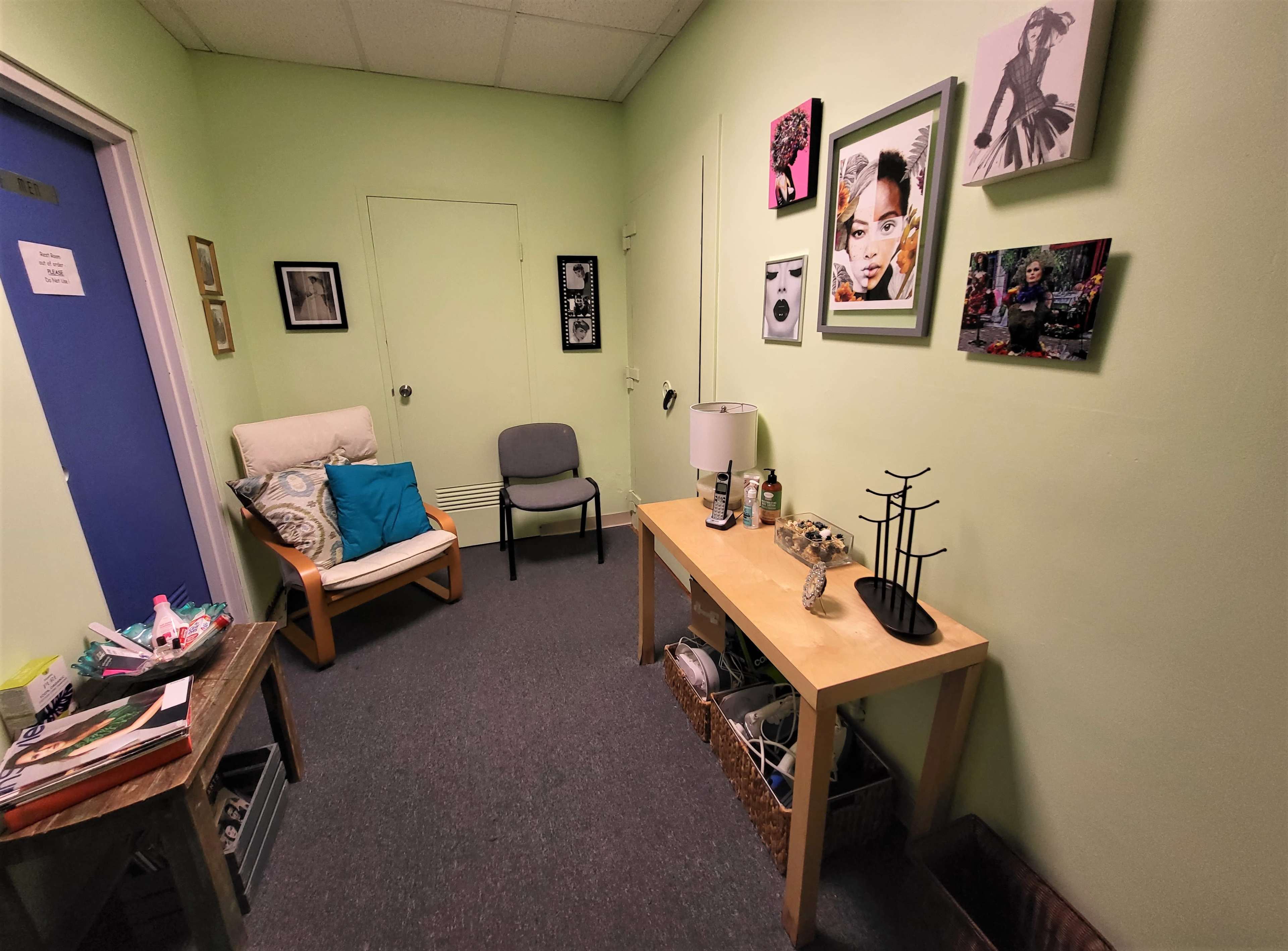 A small room with pale green walls, featuring a chair, a small table with decorative items, and framed artwork on the walls.