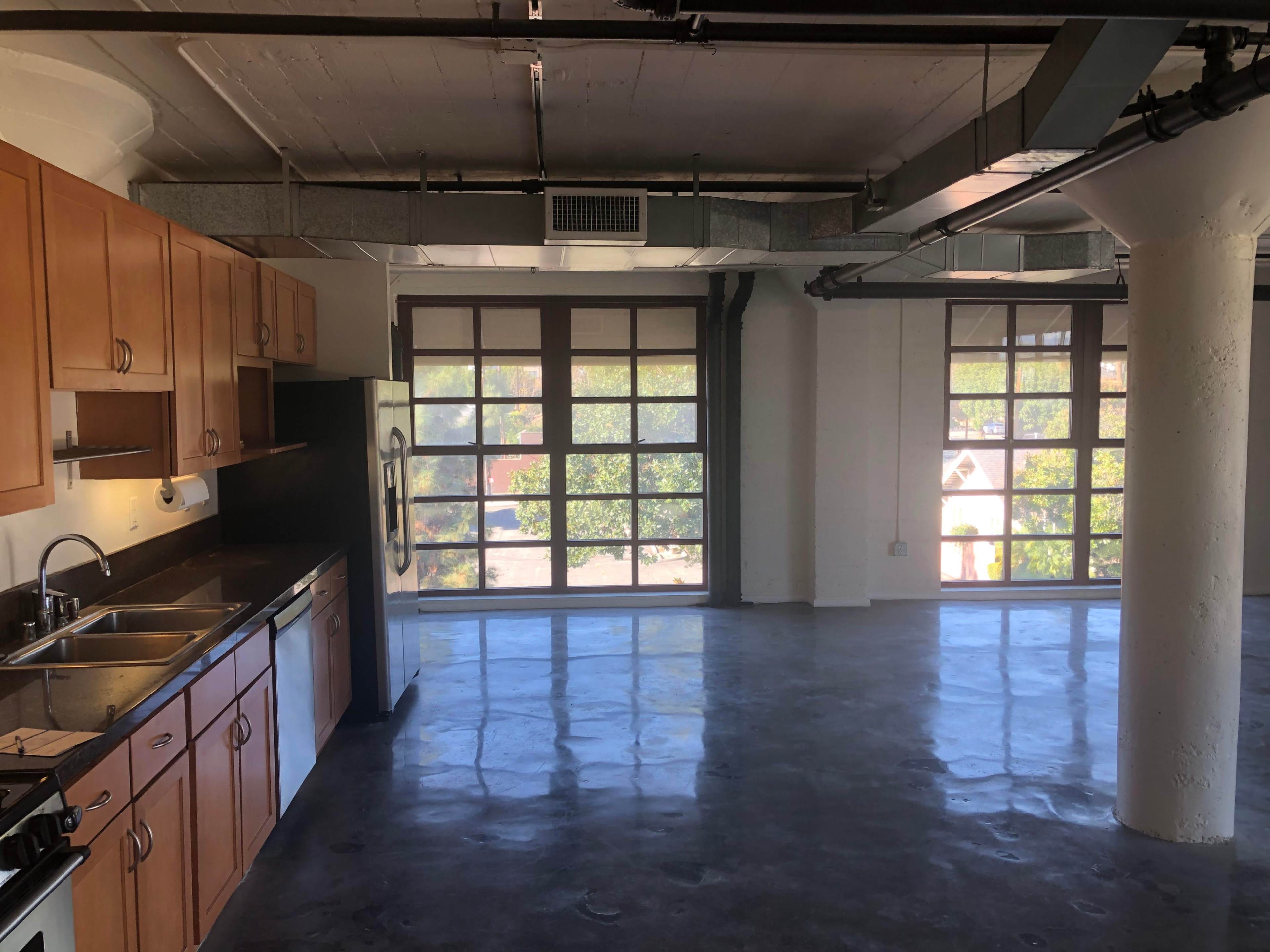 The image shows a spacious industrial kitchen area with wooden cabinets and a countertop, overlooking large windows in a modern apartment.