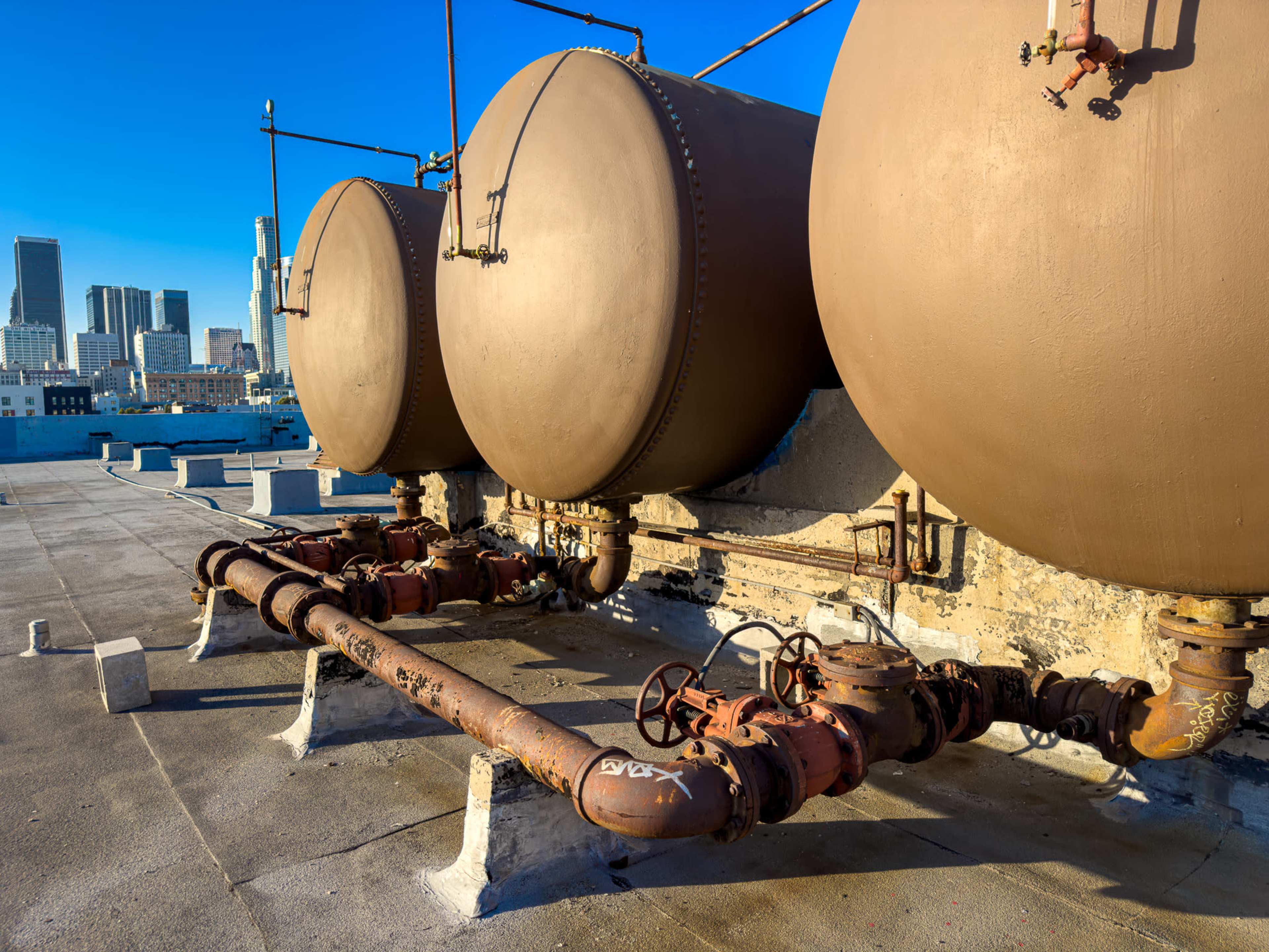 Three large, round storage tanks are mounted on a rooftop, connected by rusted pipes and valves, with a city skyline in the background.