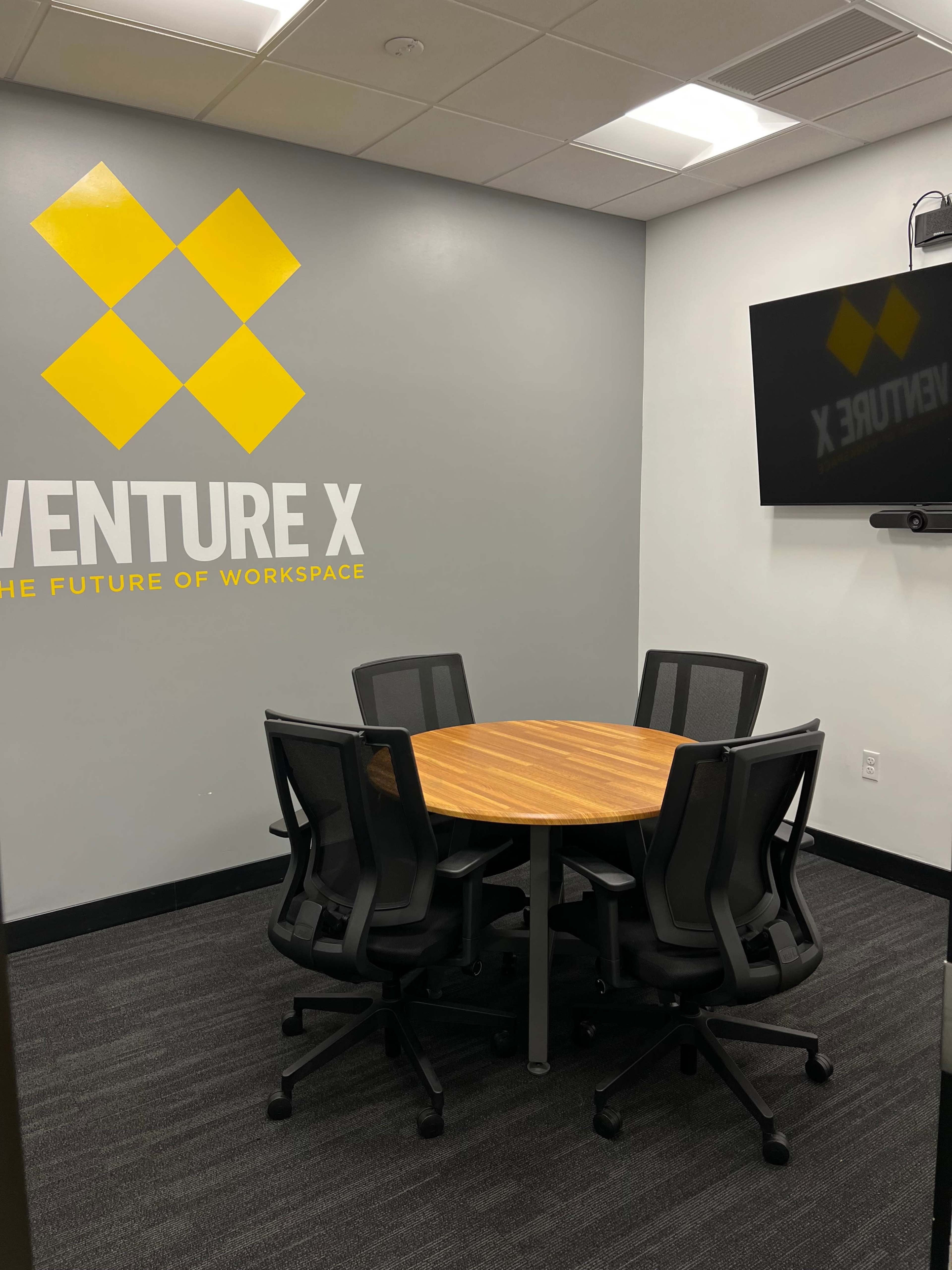 A small conference room features a round wooden table surrounded by six black chairs, with a wall displaying the "Venture X" logo.