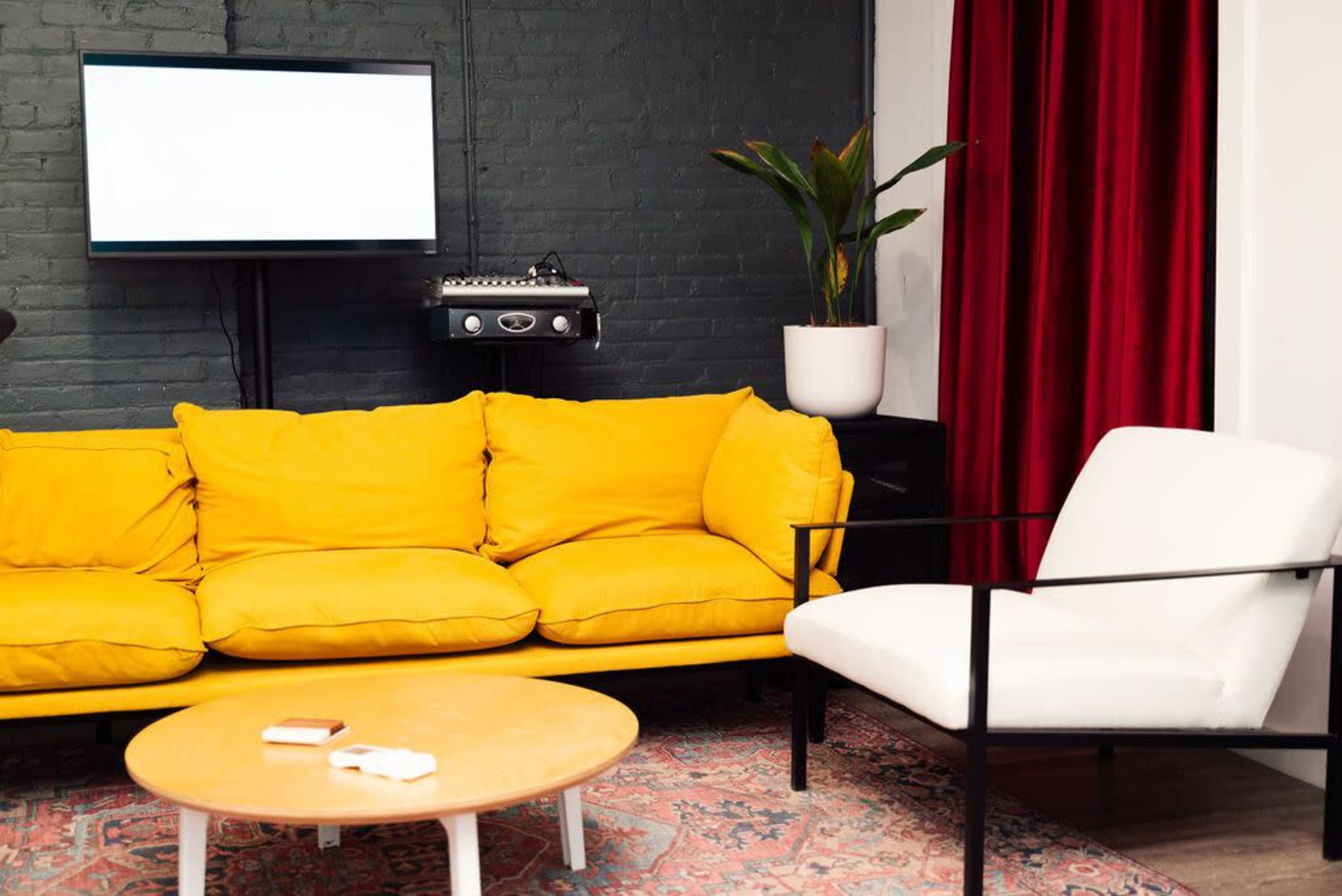 A bright yellow couch and a white armchair are positioned in a room with green brick walls, a red curtain, and a plant beside a wall-mounted TV.