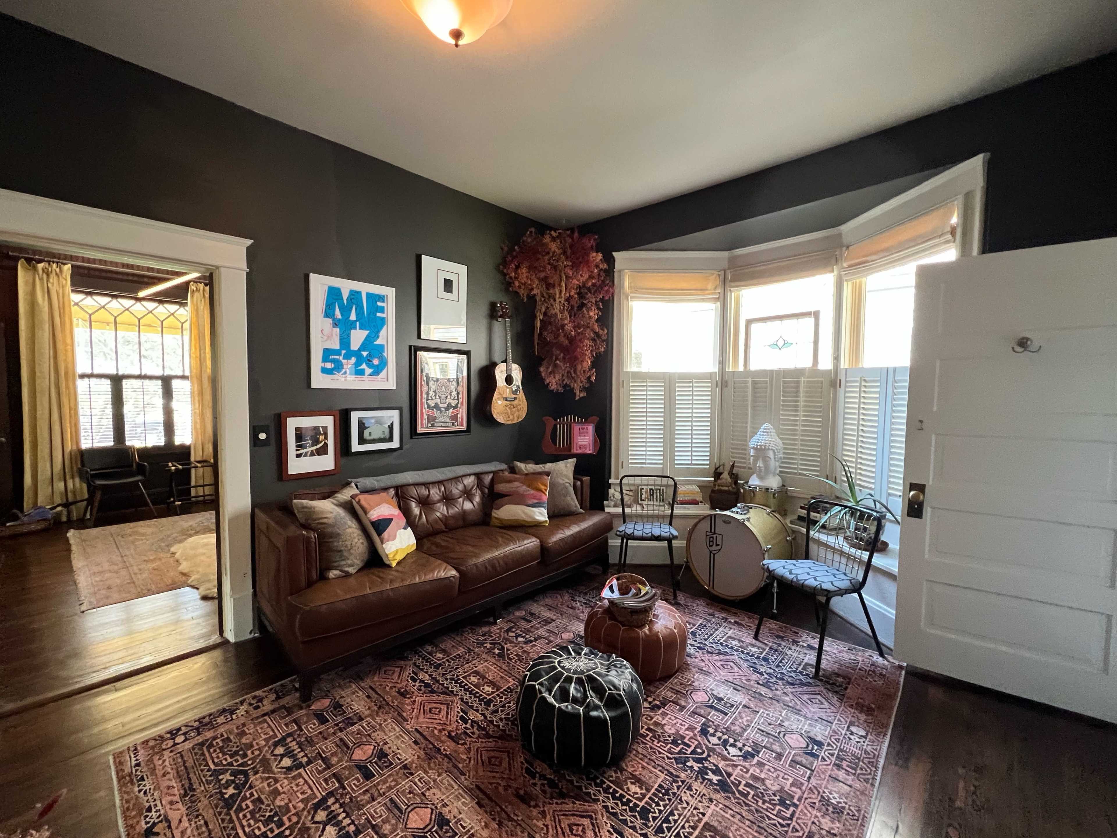 A cozy living room with a brown leather sofa, various framed artworks on the walls, and a large area rug under a round ottoman and decorative chairs near a bay window.