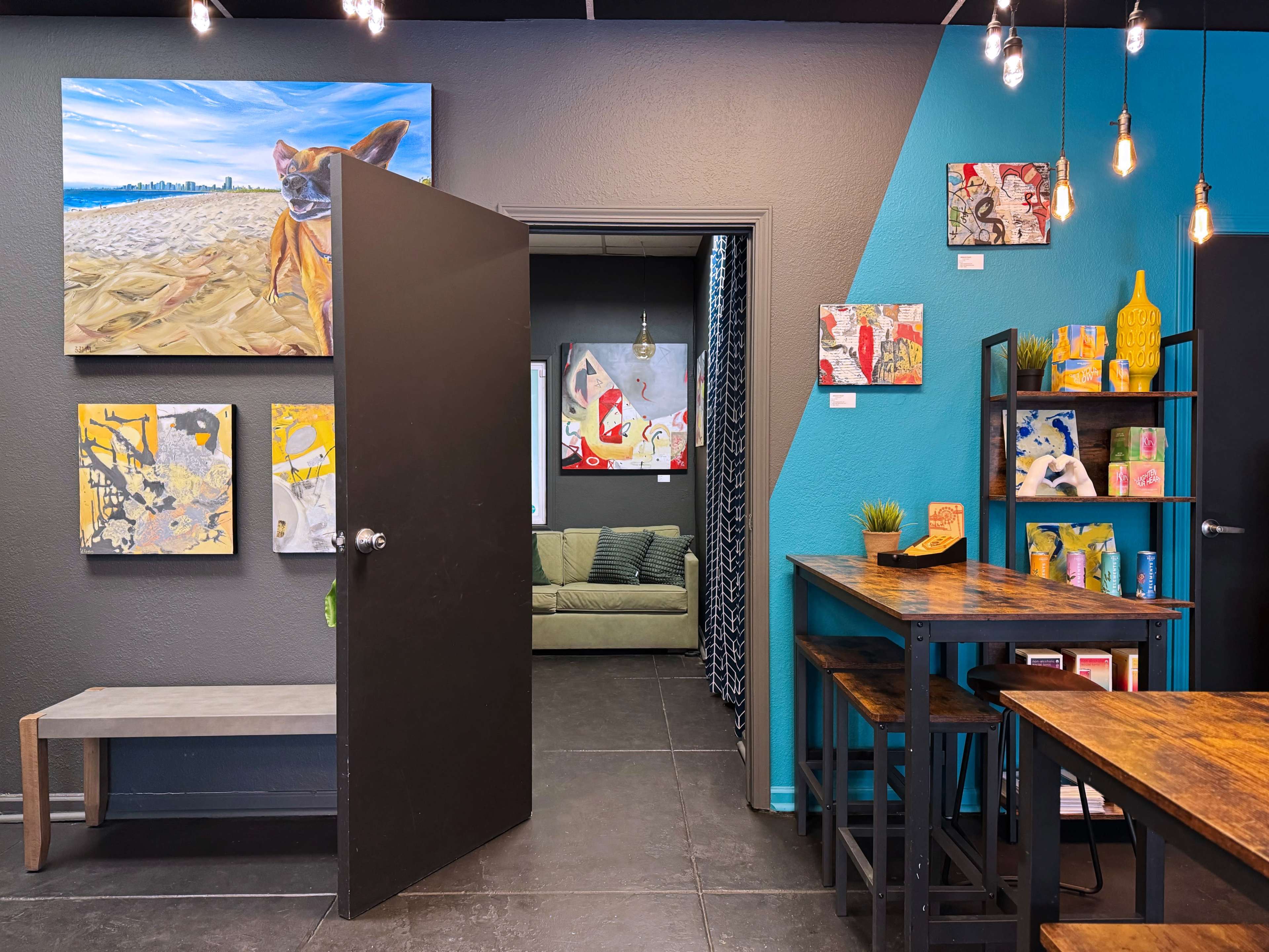 An interior space featuring colorful artwork on the walls, a bench, a green couch, and a dining area with wooden tables and stools.
