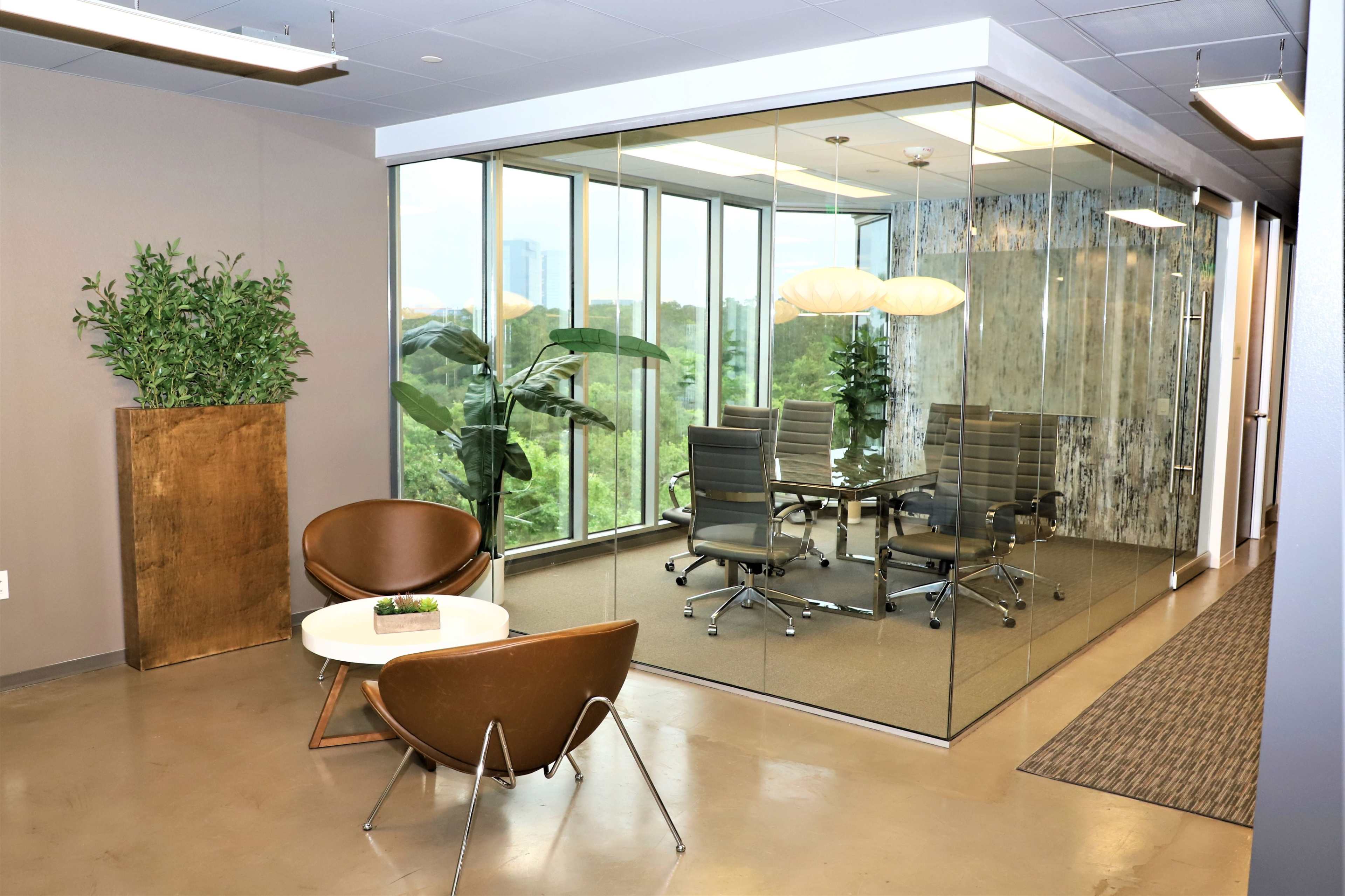 A modern office space features a glass-walled meeting room with a conference table and ergonomic chairs, surrounded by plants and soft lighting.