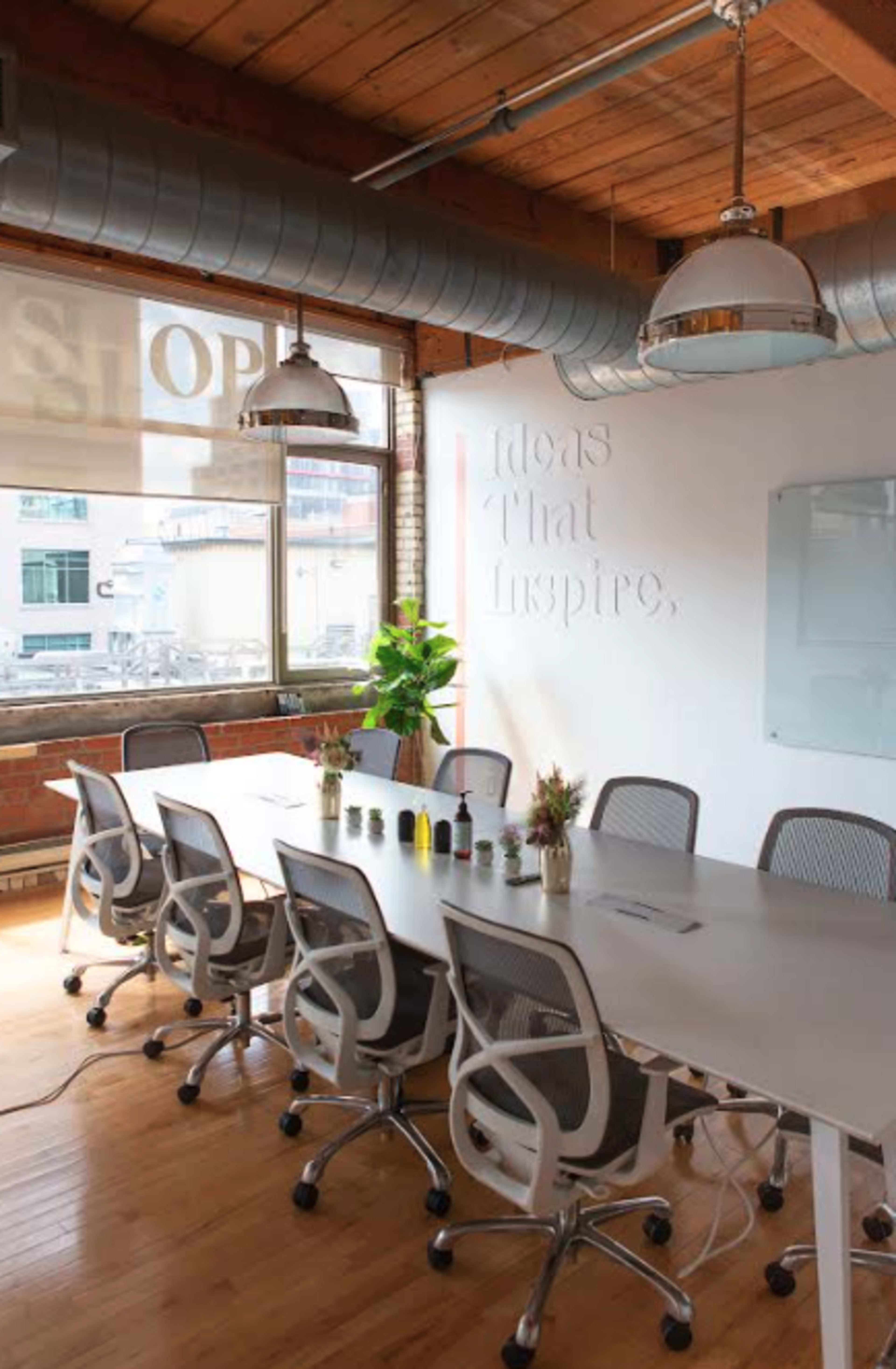 A modern conference room features a long white table surrounded by ergonomic chairs, with large windows allowing natural light and a wall displaying the phrase "Ideas That Inspire."