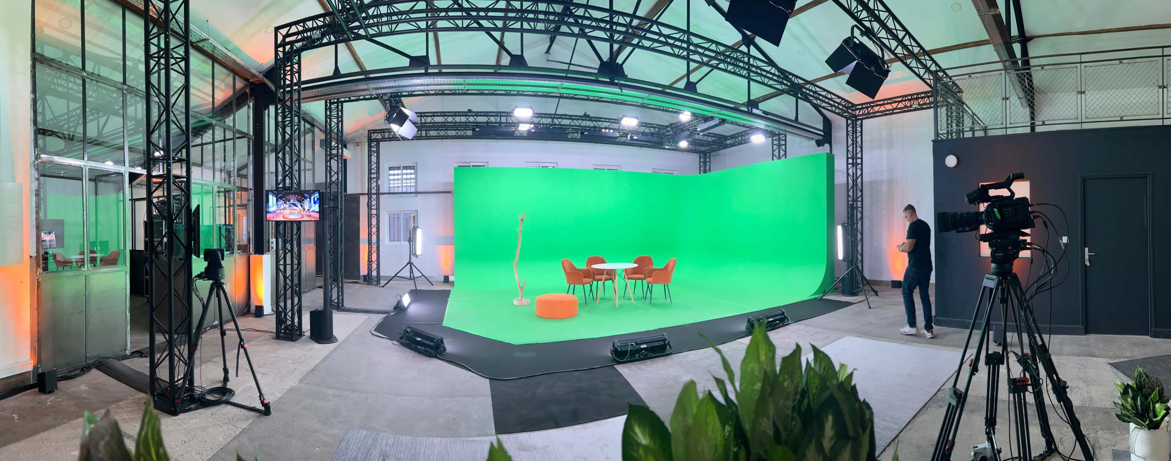 A spacious studio with a green screen backdrop, a circular table surrounded by four orange chairs, and studio lighting equipment set up for a production.