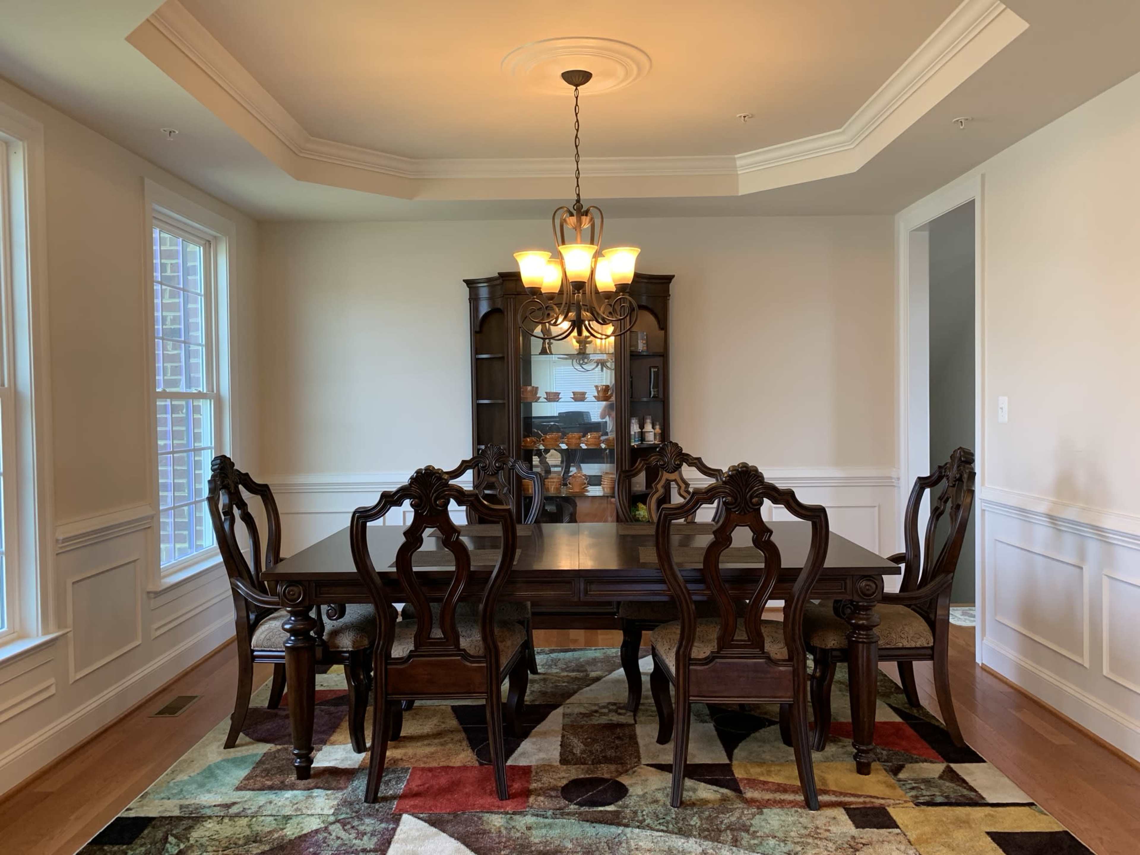 A dining room with a dark wooden table surrounded by six ornate chairs, illuminated by a chandelier, with a cabinet displaying items in the background.