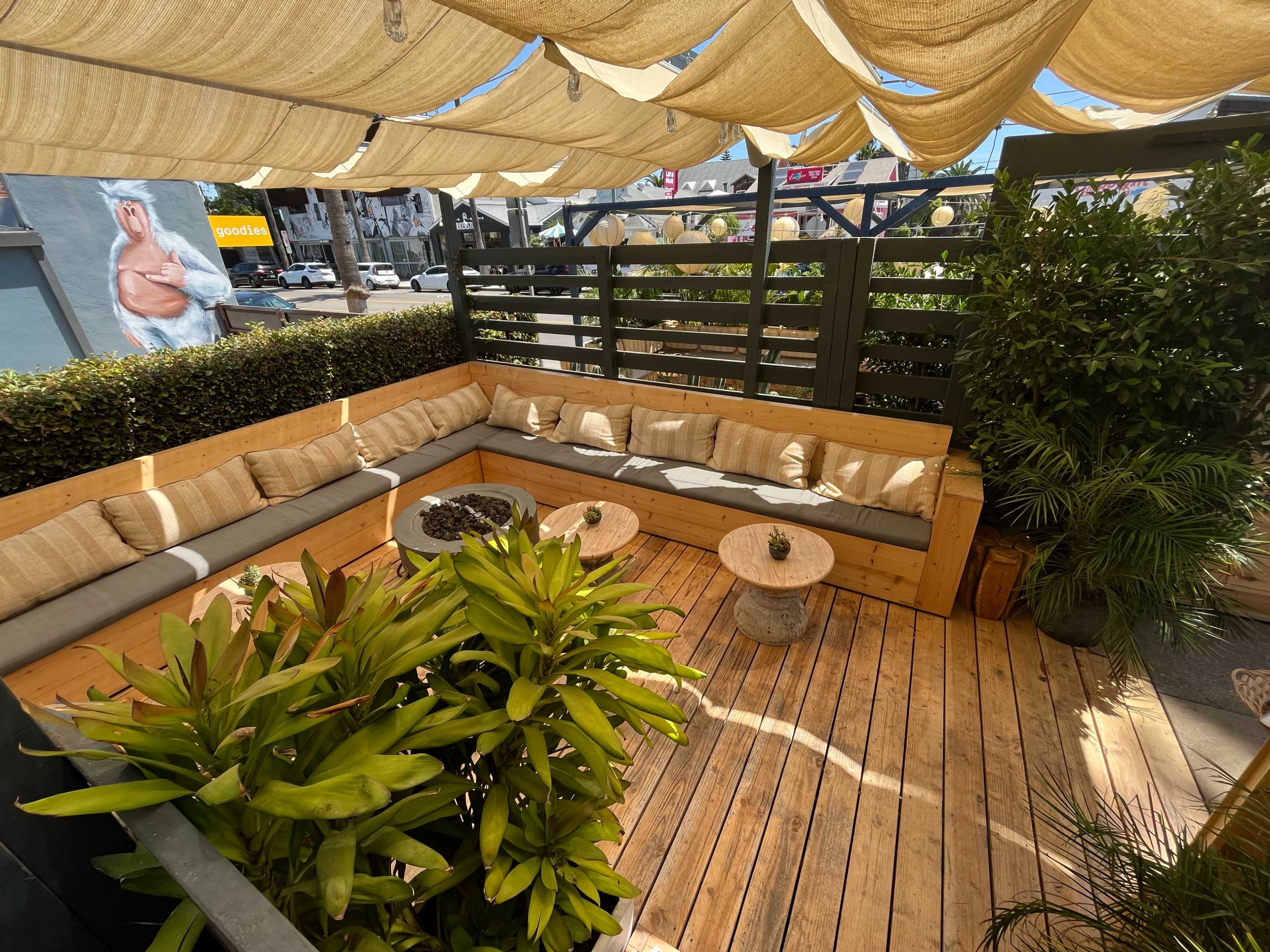 A wooden outdoor seating area is enclosed by low greenery and features a shaded couch with cushions, two small round tables, and potted plants.