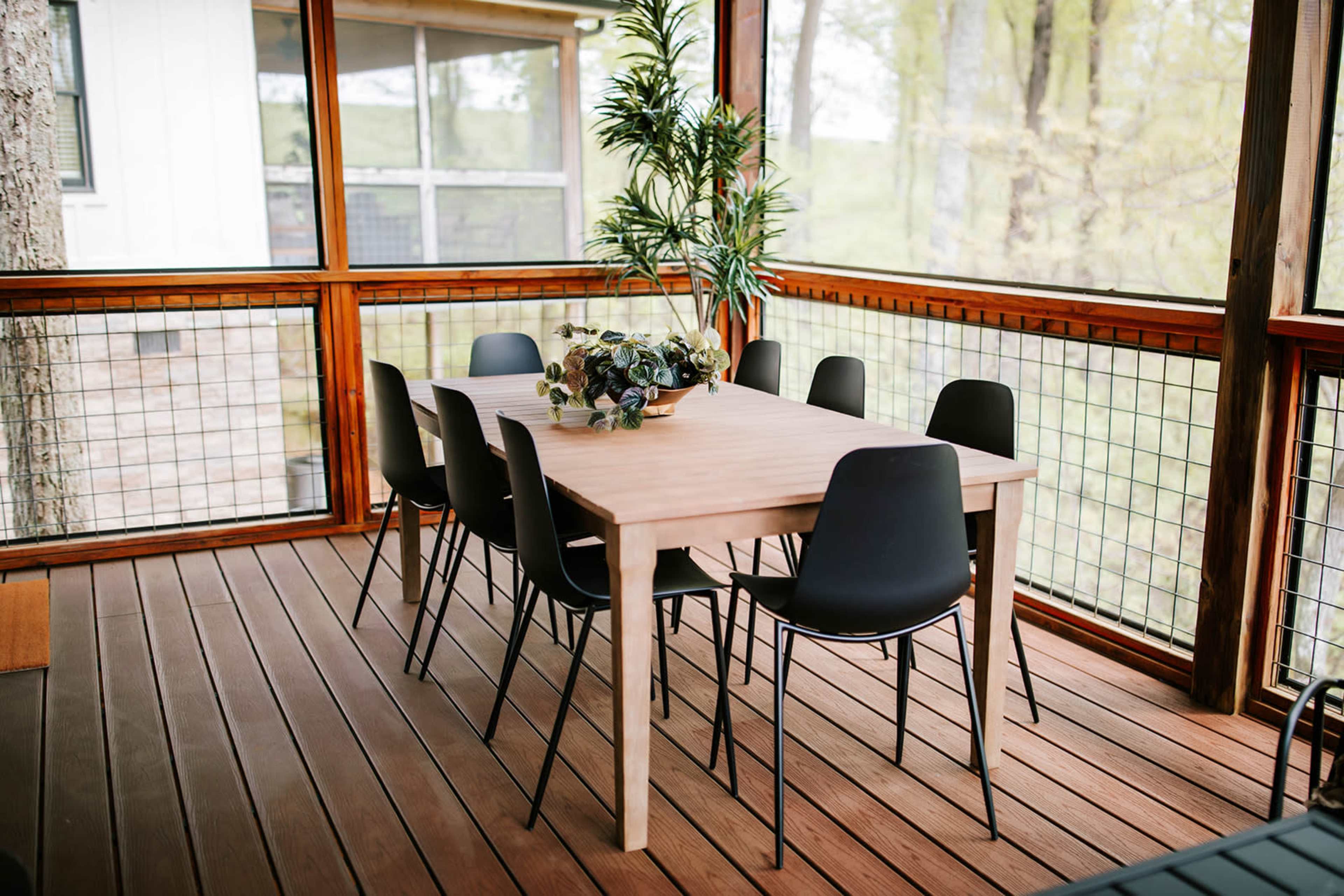 A wooden dining table with eight black chairs is set on a wooden deck surrounded by large windows and greenery.