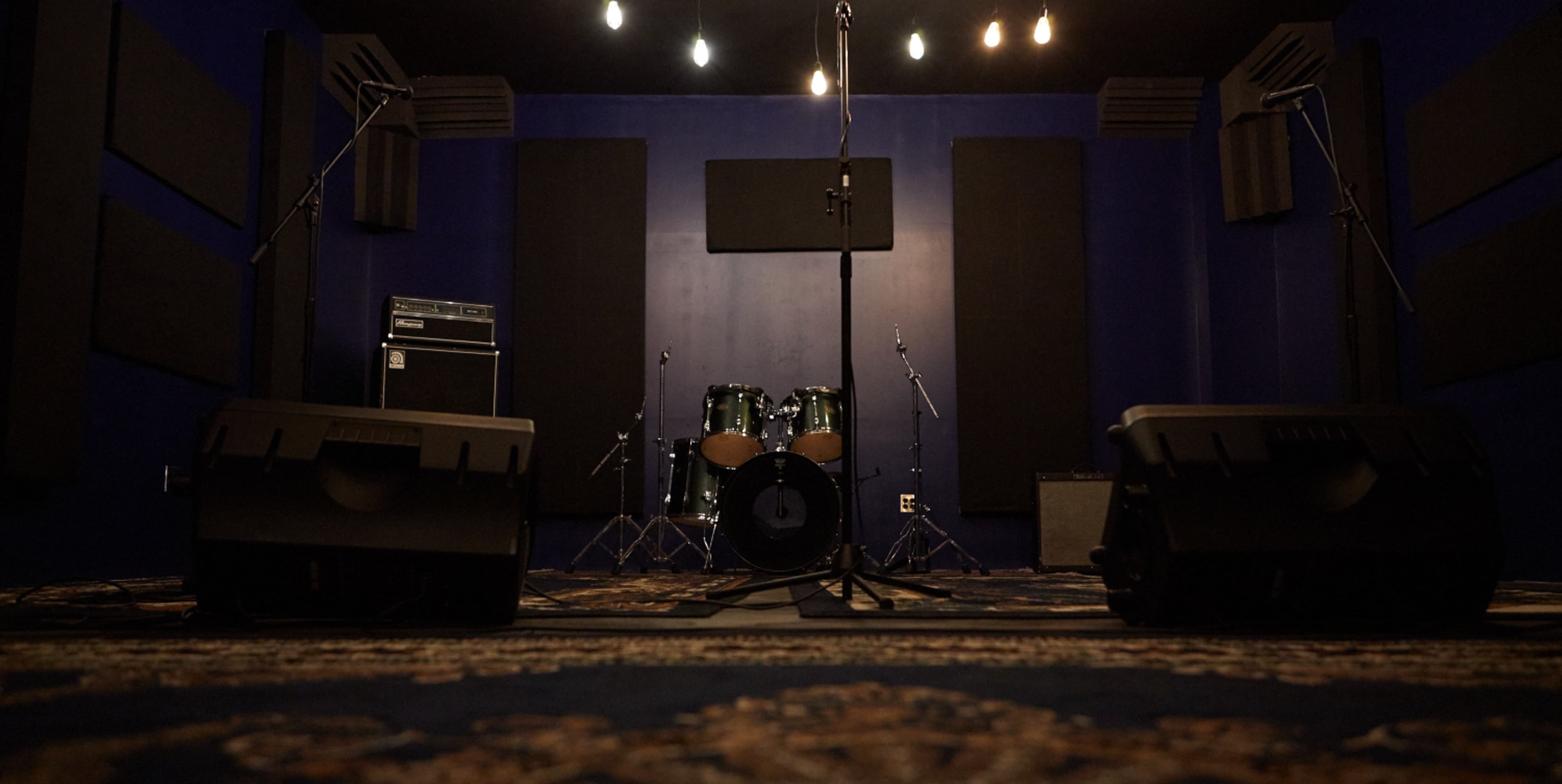 The image shows a music rehearsal space with a drum set and microphones set up on stage, surrounded by soundproof panels.