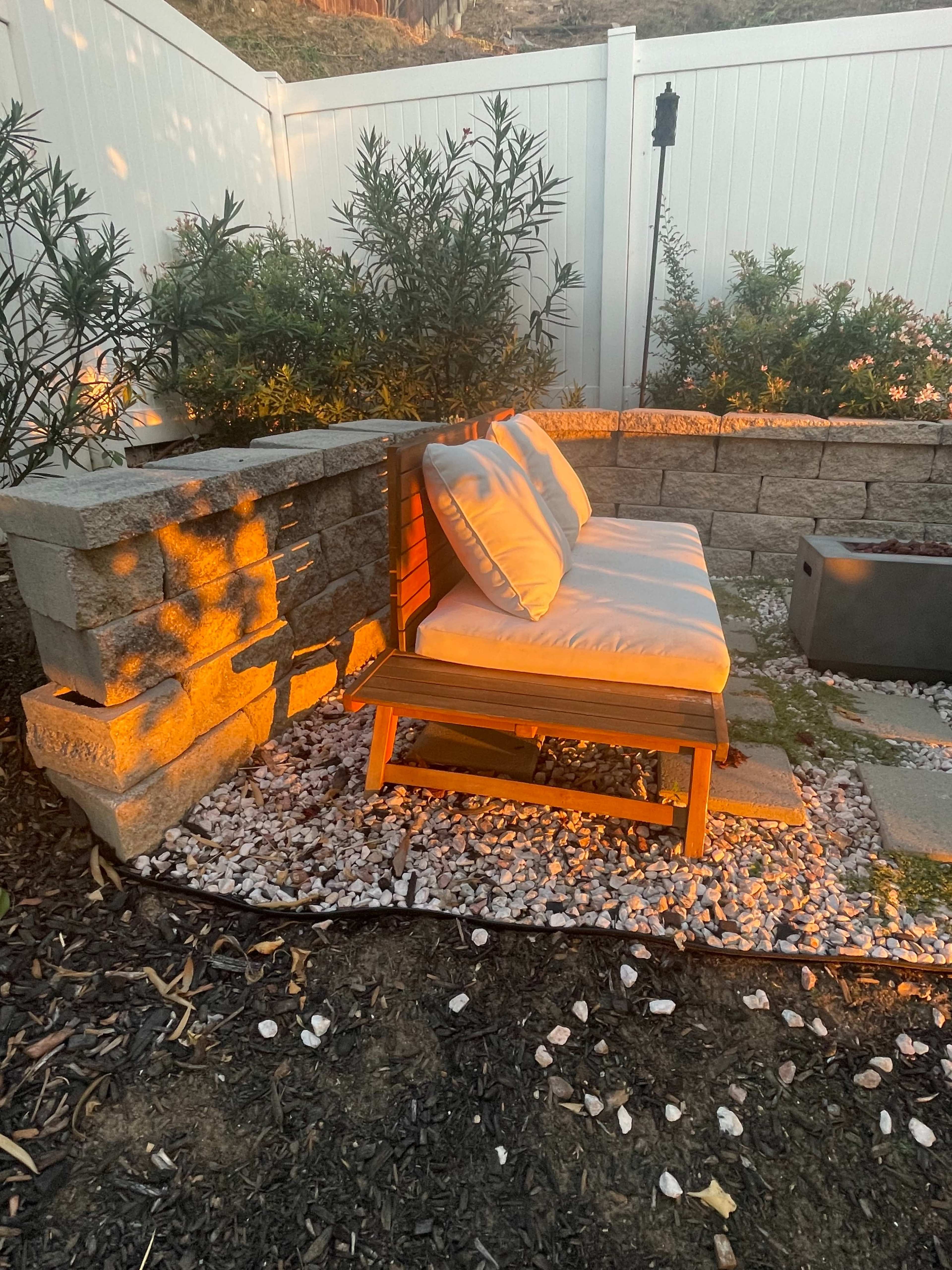 A wooden bench with white cushions is positioned beside a stone wall in a landscaped outdoor area.