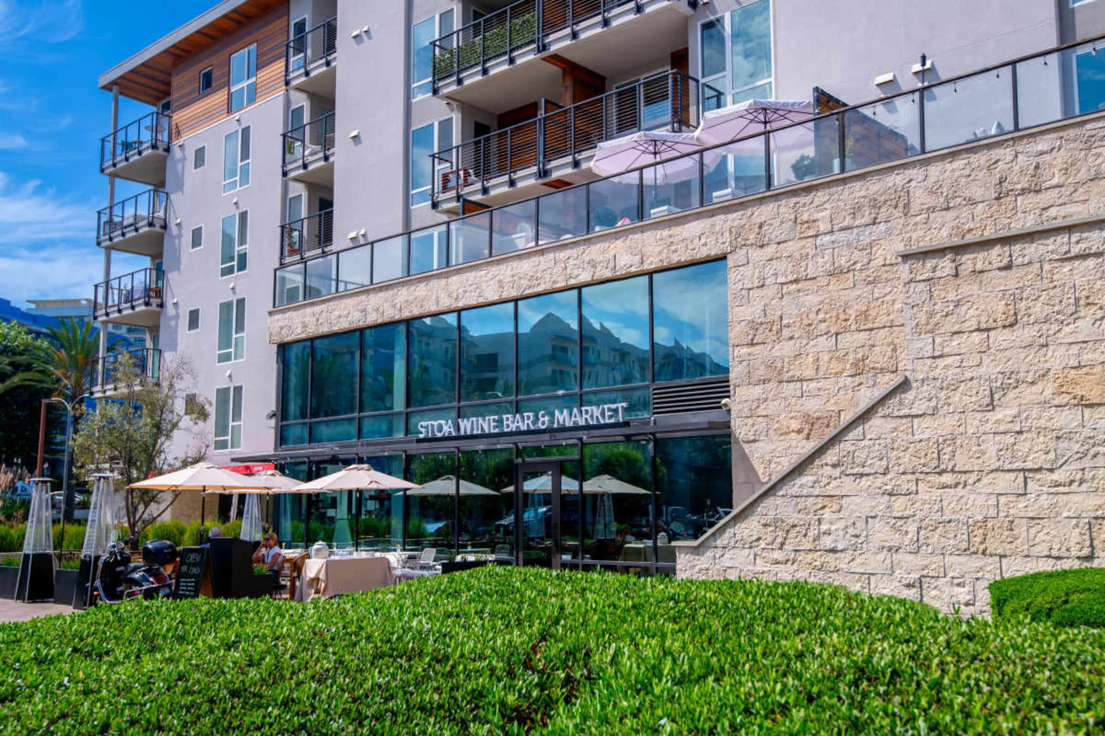 The image shows a building with a glass facade that houses a store named "Stock Wine Bar & Market," surrounded by greenery and outdoor seating areas.