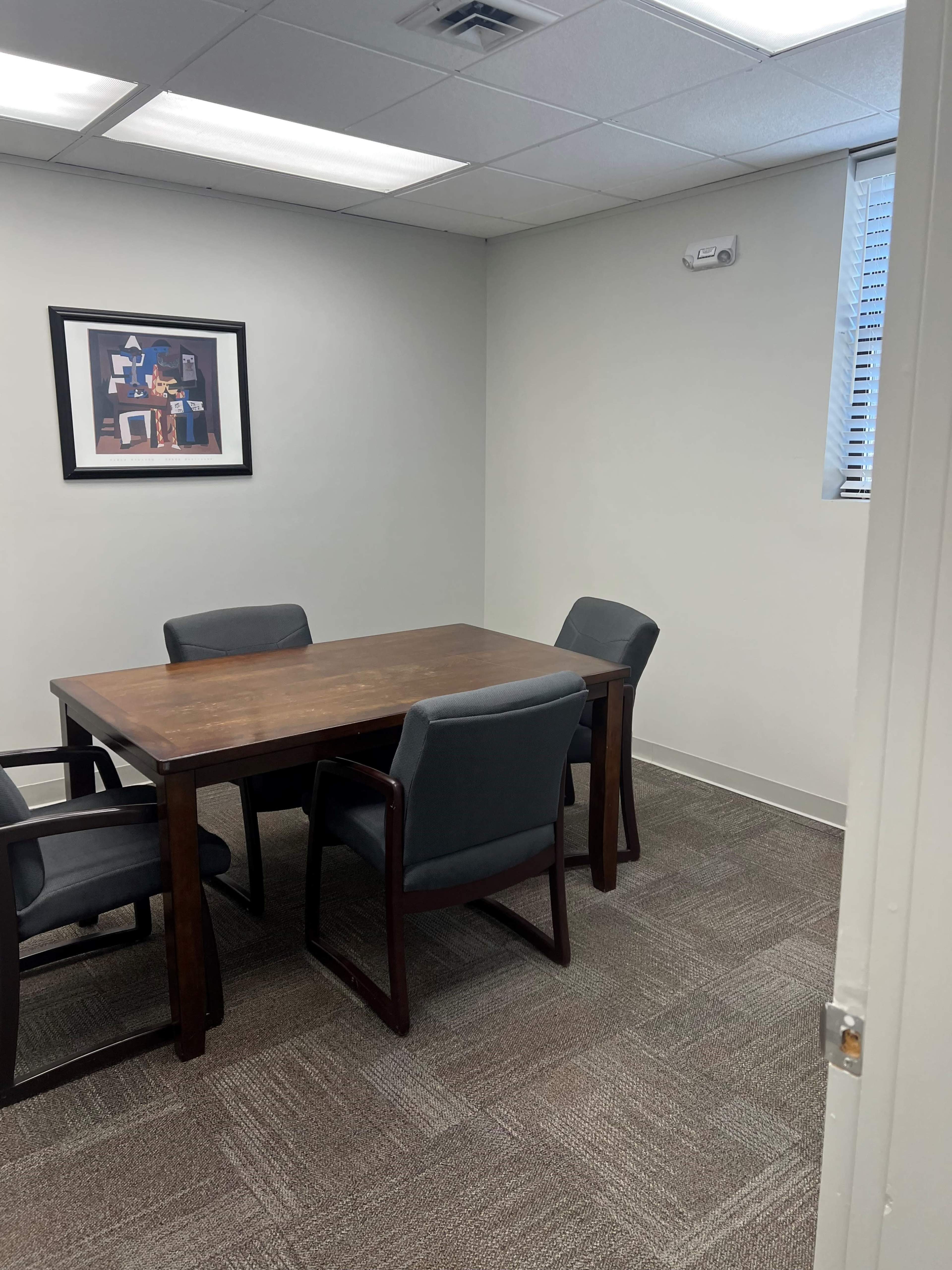 A wooden table with four chairs is placed in a small, well-lit conference room featuring a framed art piece on the wall.