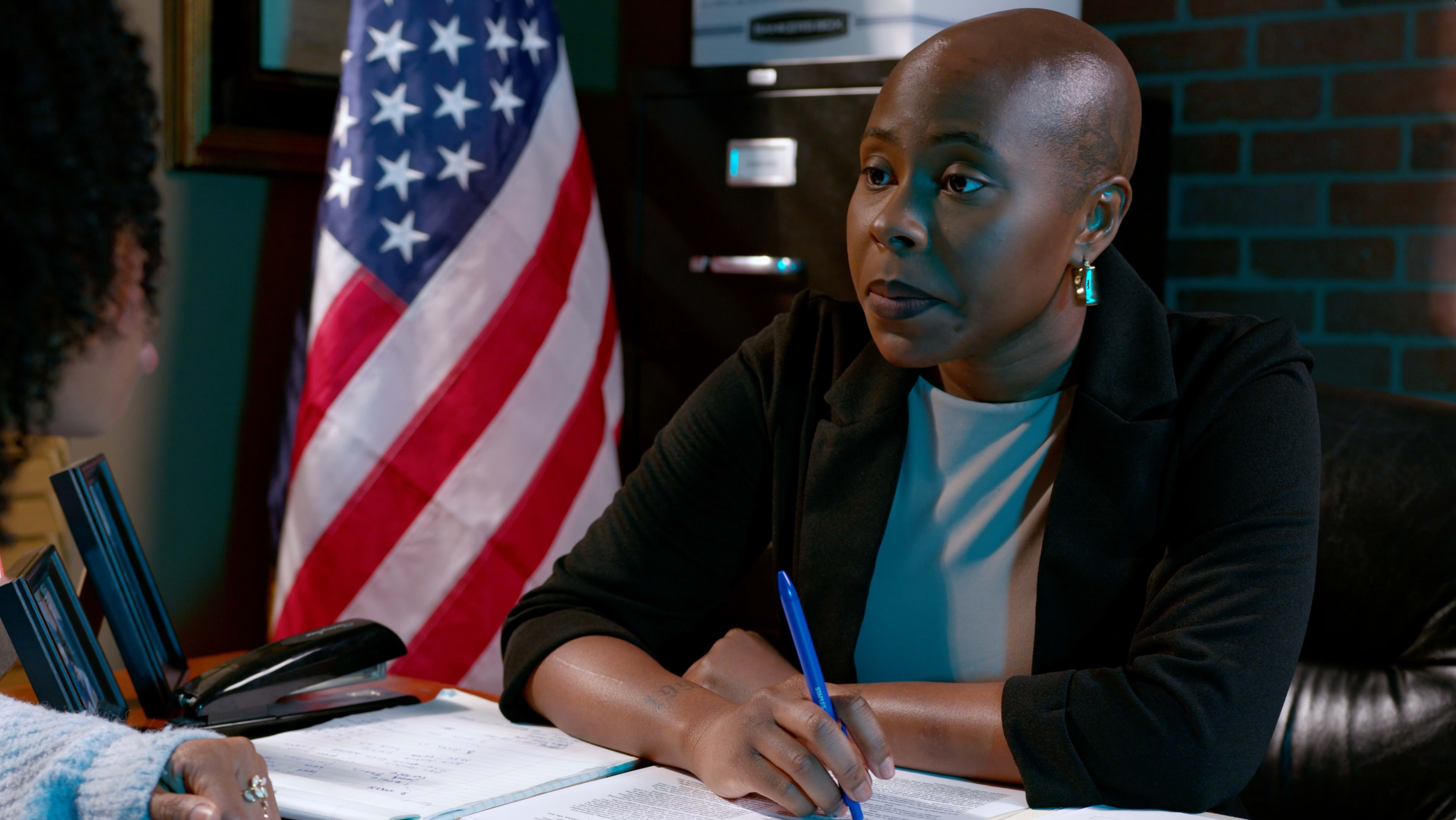 A woman with a bald head sits at a desk with an American flag in the background, writing notes during a conversation.