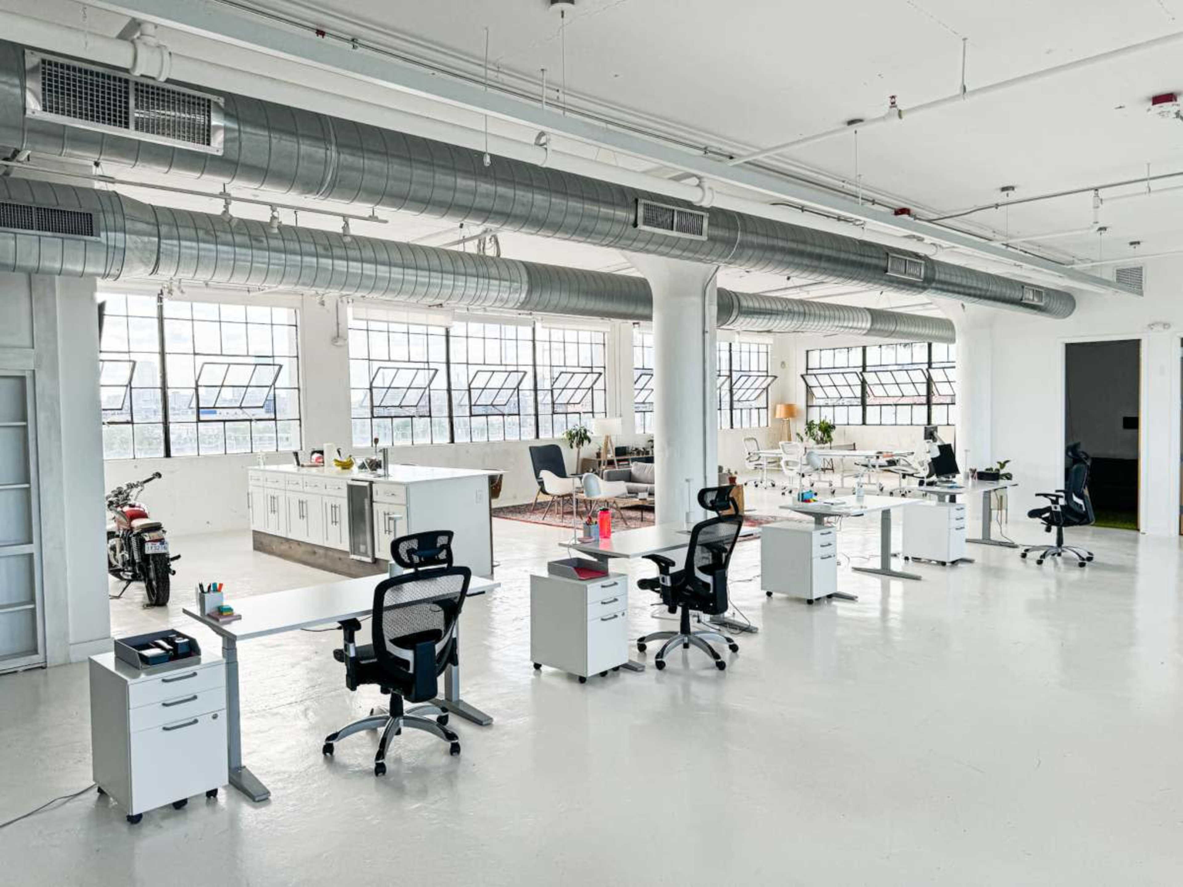 The image shows a spacious modern office with several desks, ergonomic chairs, and large windows, along with a kitchenette in the background.