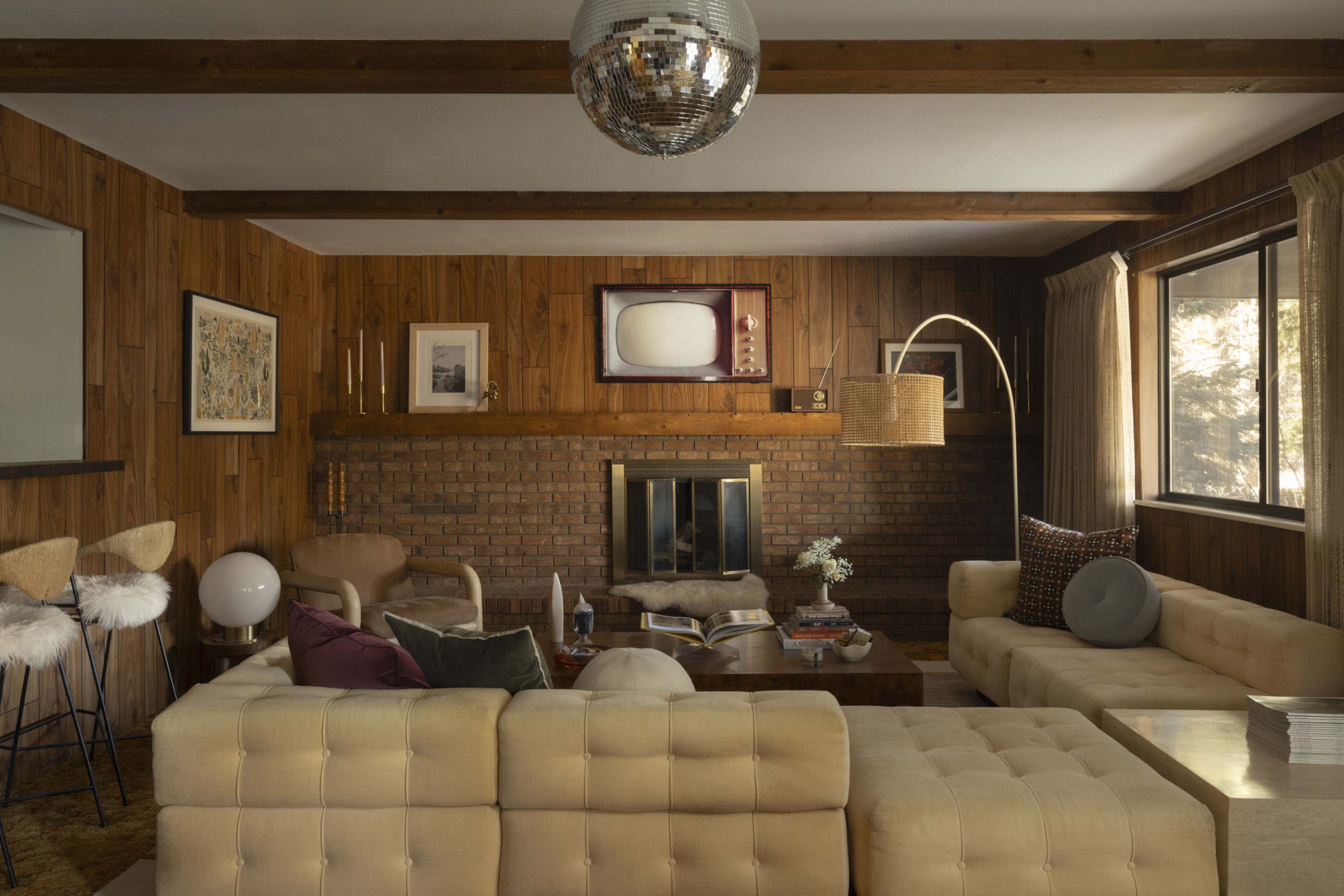 The image shows a mid-century modern living room with wooden walls, a brick fireplace, and a mix of vintage furniture, including a white tufted sofa and an old television on the wall.