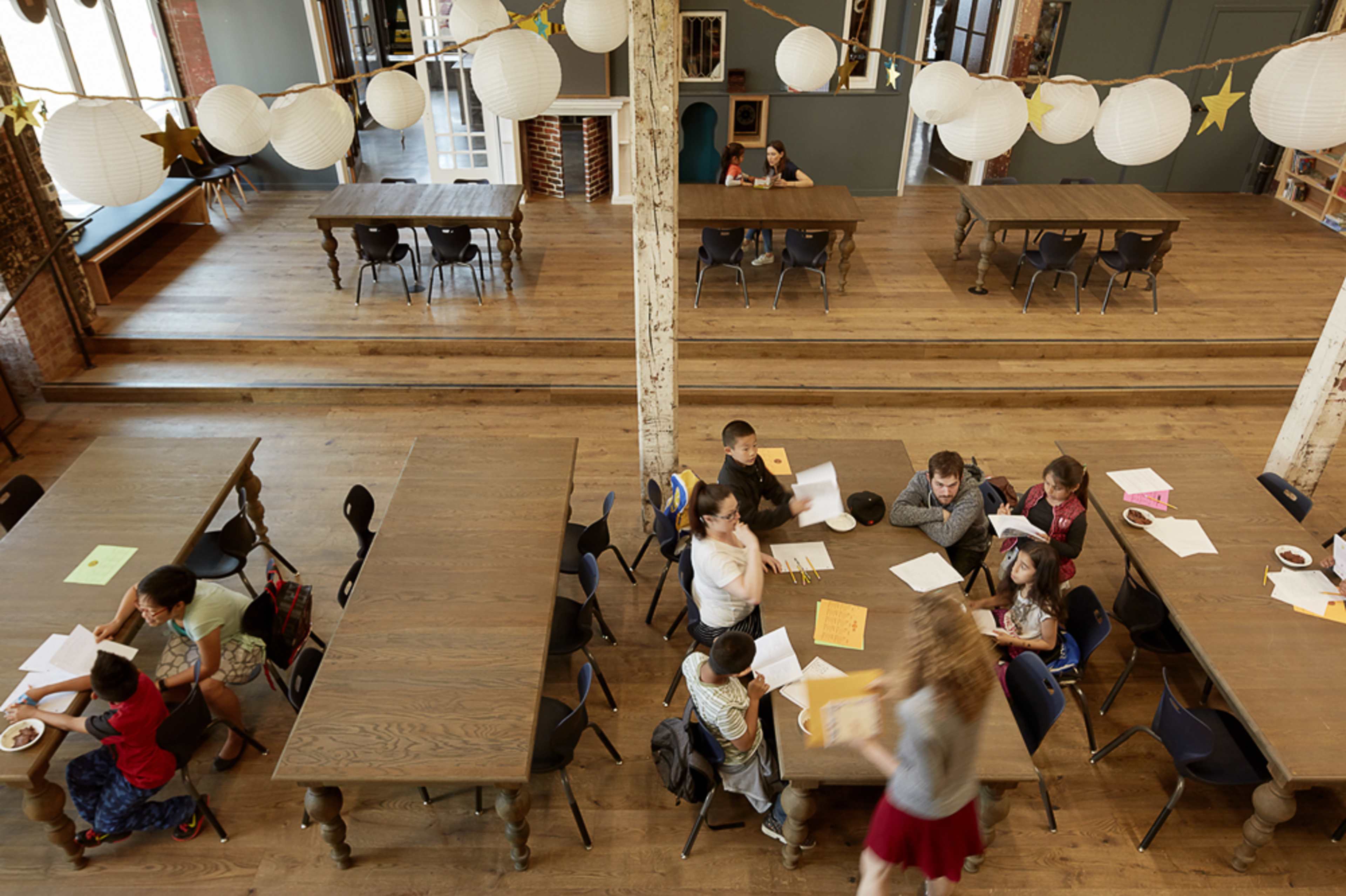 A spacious room with several wooden tables where children and adults are engaged in activities, with paper and food items scattered on the tables.