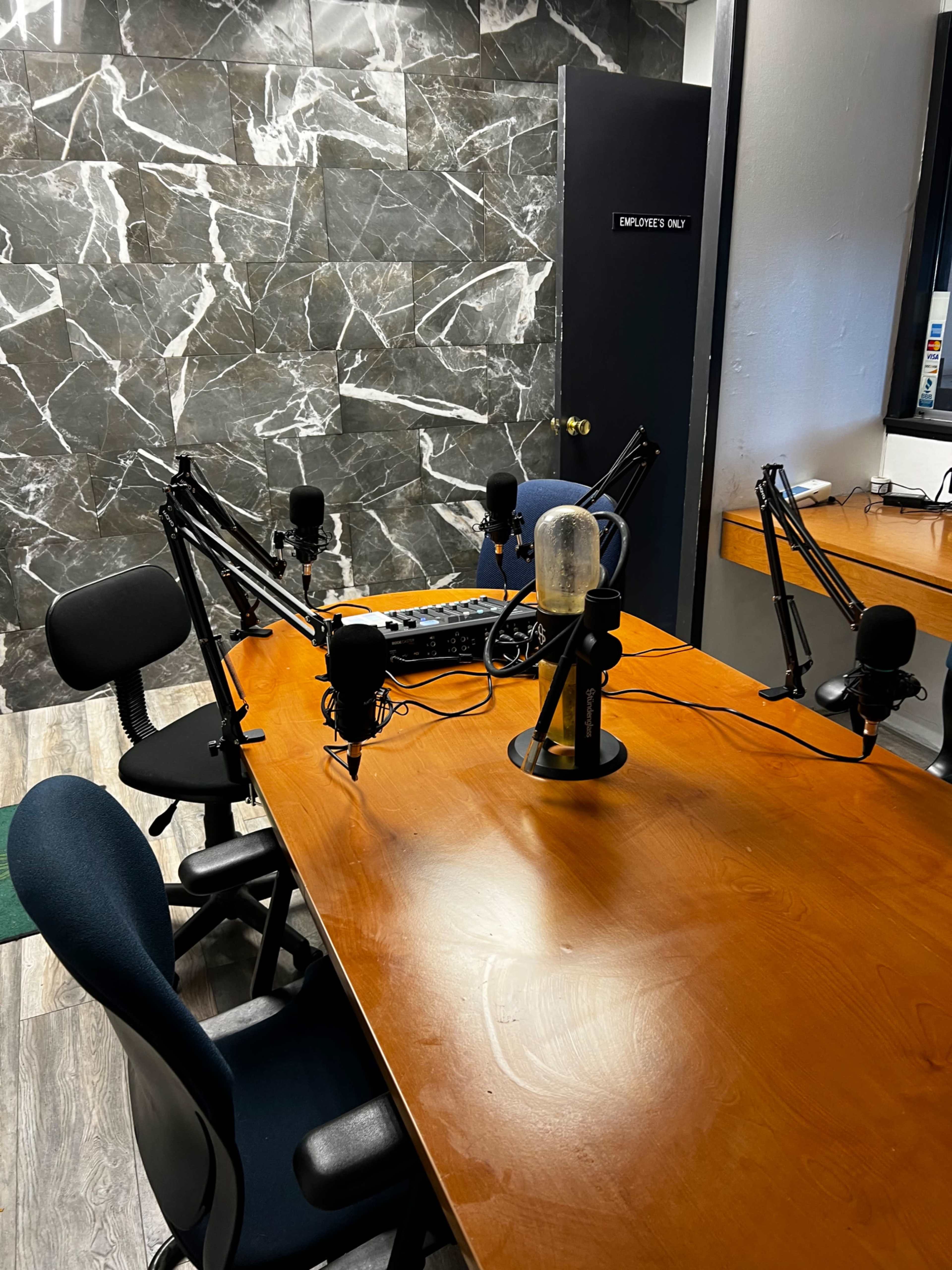 A podcast studio featuring a wooden table with several microphones mounted on stands and a soundboard, against a marble-patterned wall.