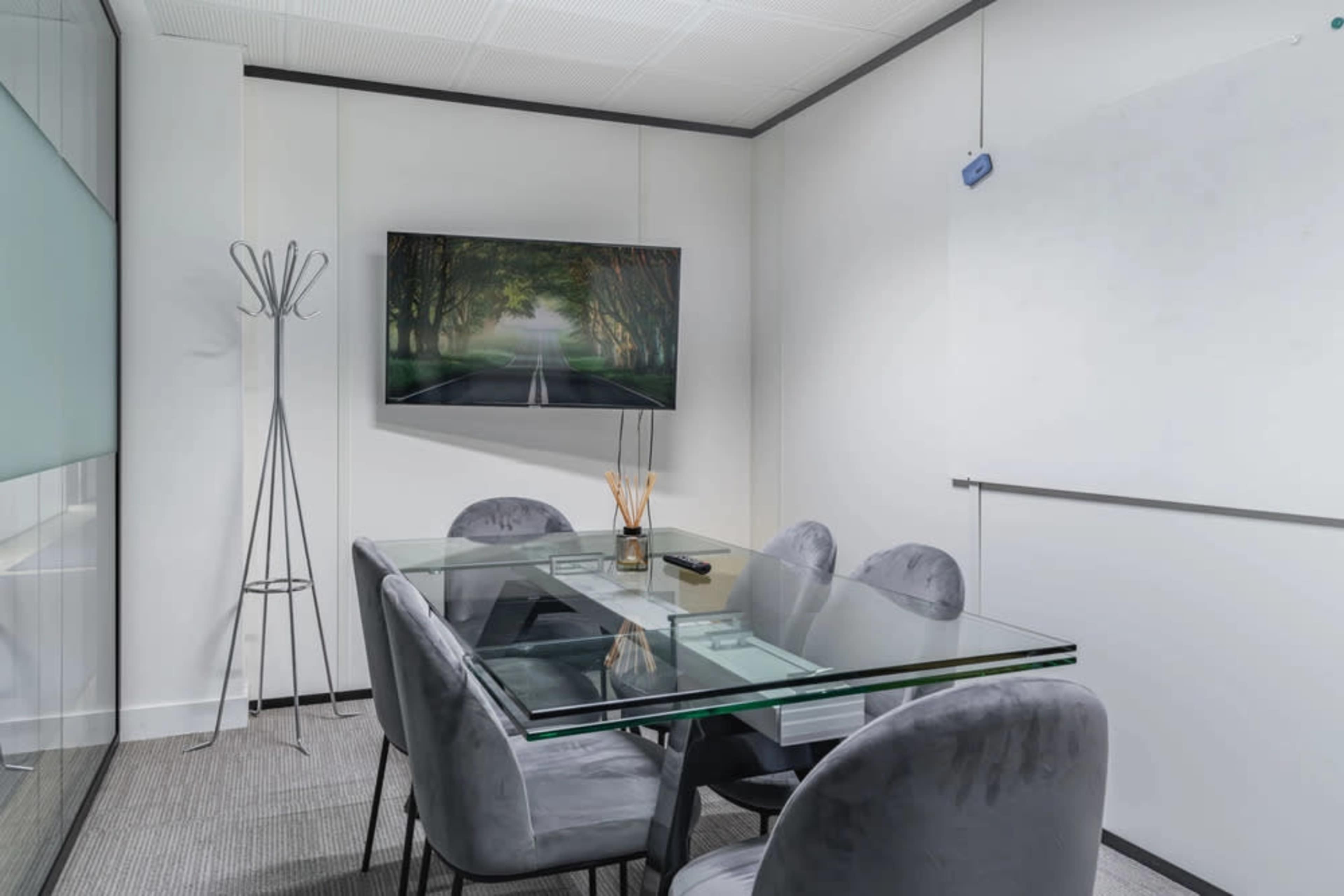 The image shows a modern conference room with a glass table, four upholstered chairs, a wall-mounted television, and a coat stand.