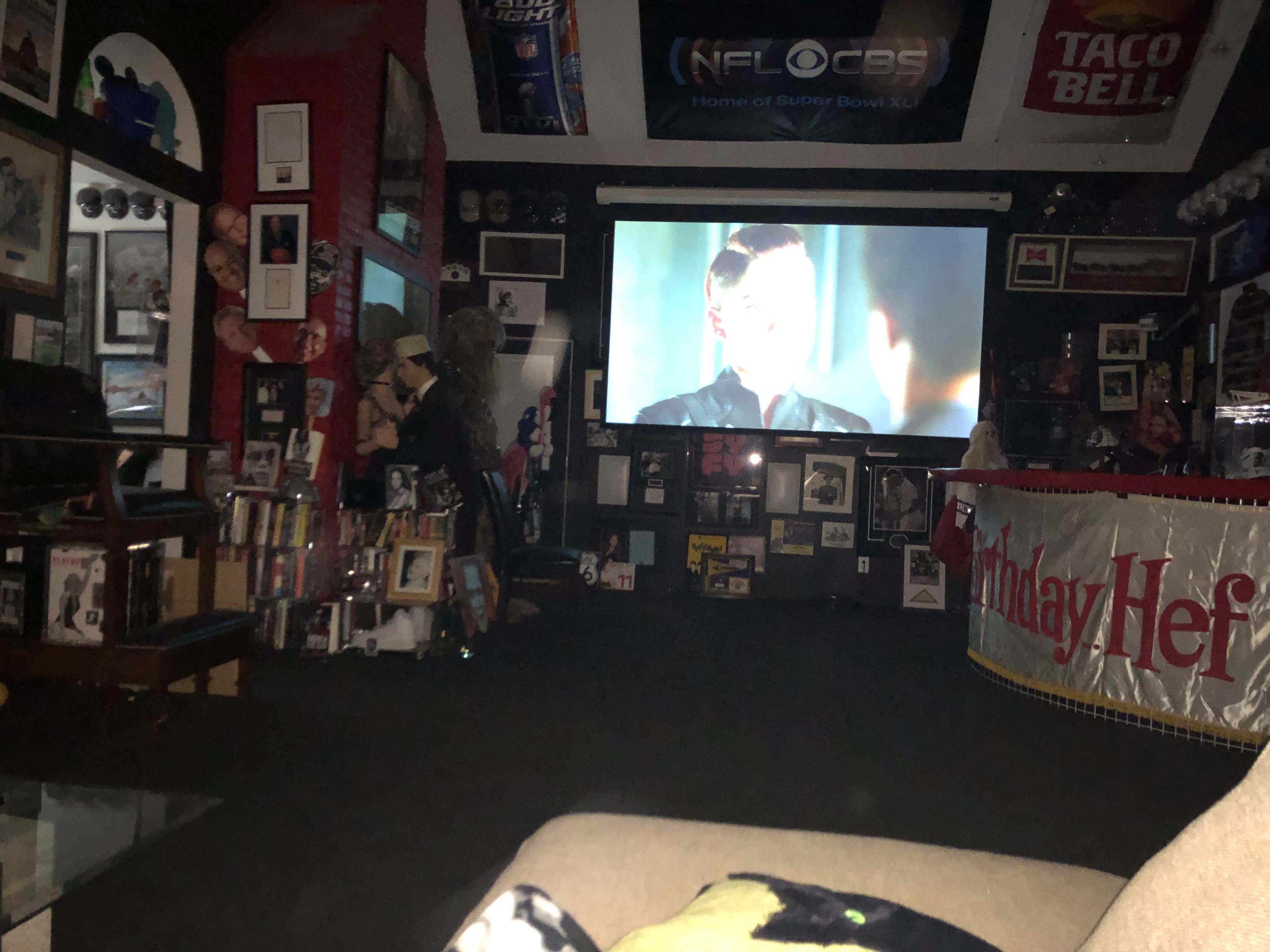 The image shows a room with a large screen displaying a scene from a movie or television show, surrounded by various framed photographs and memorabilia.