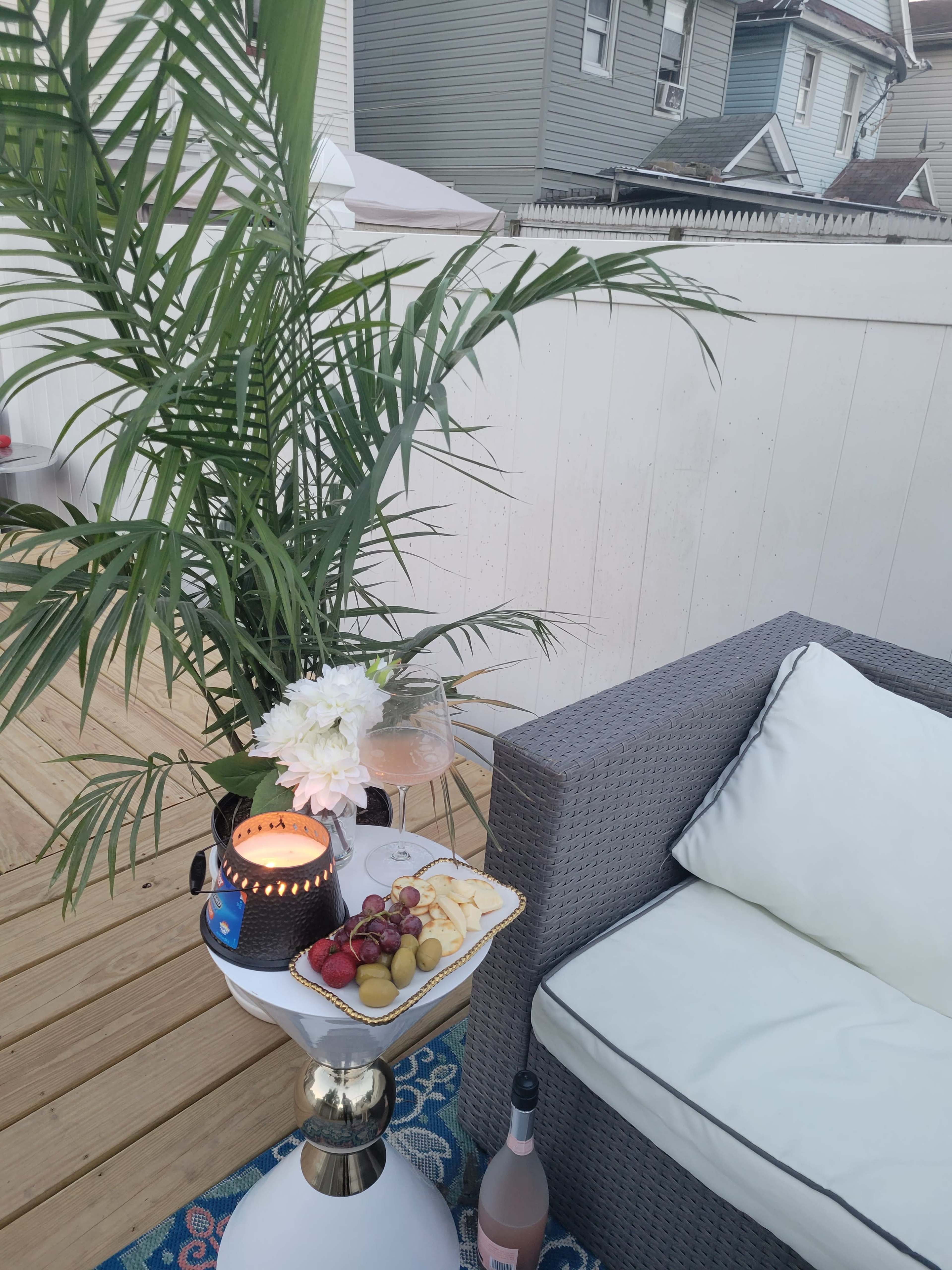 A cozy outdoor seating area features a small table with a candle, fruit platter, and a bottle of rosé, surrounded by a potted palm and a comfortable couch.