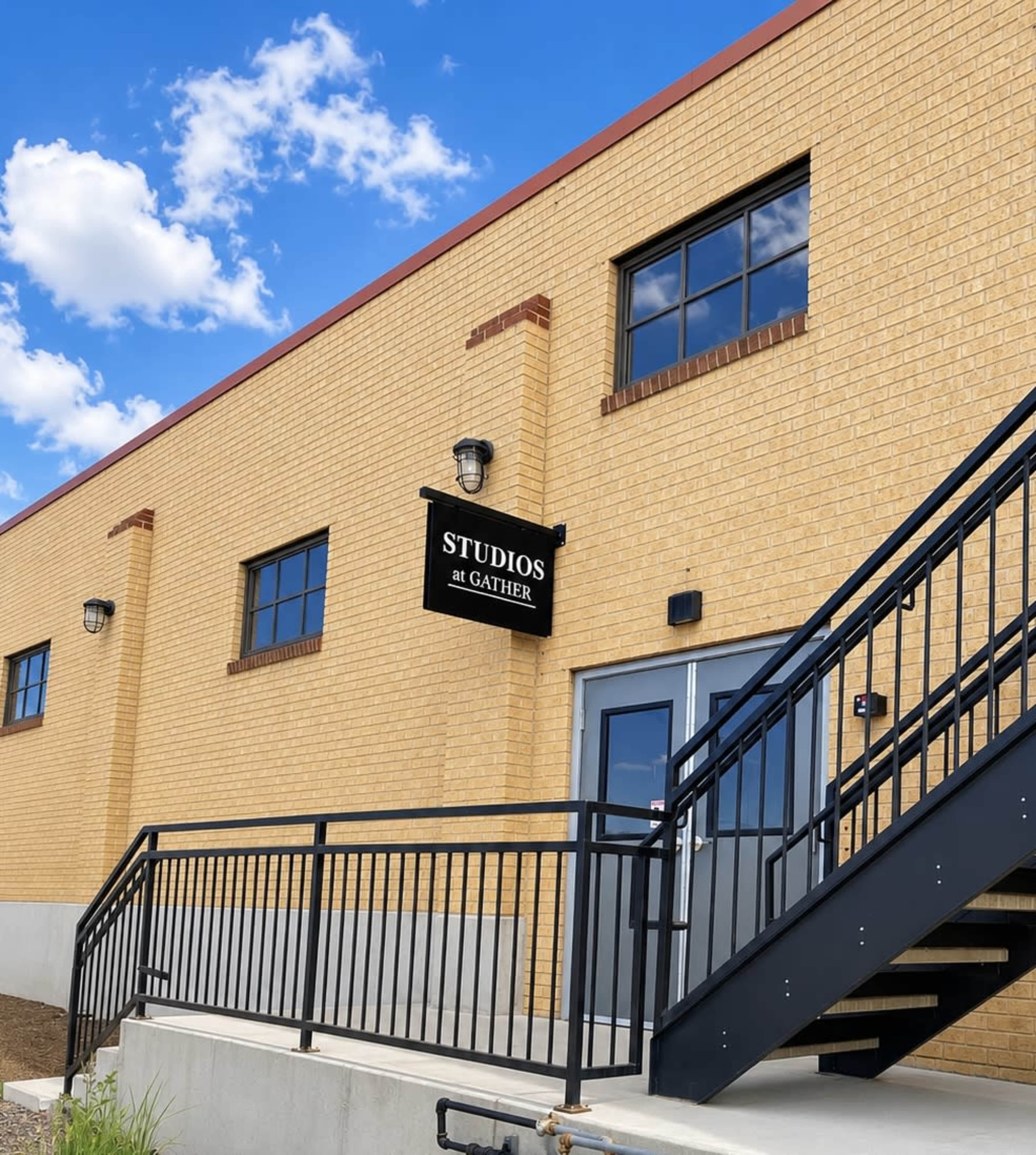 A tan-brick building features a set of metal stairs leading to a door with a sign that reads "STUDIOS at GATHER."