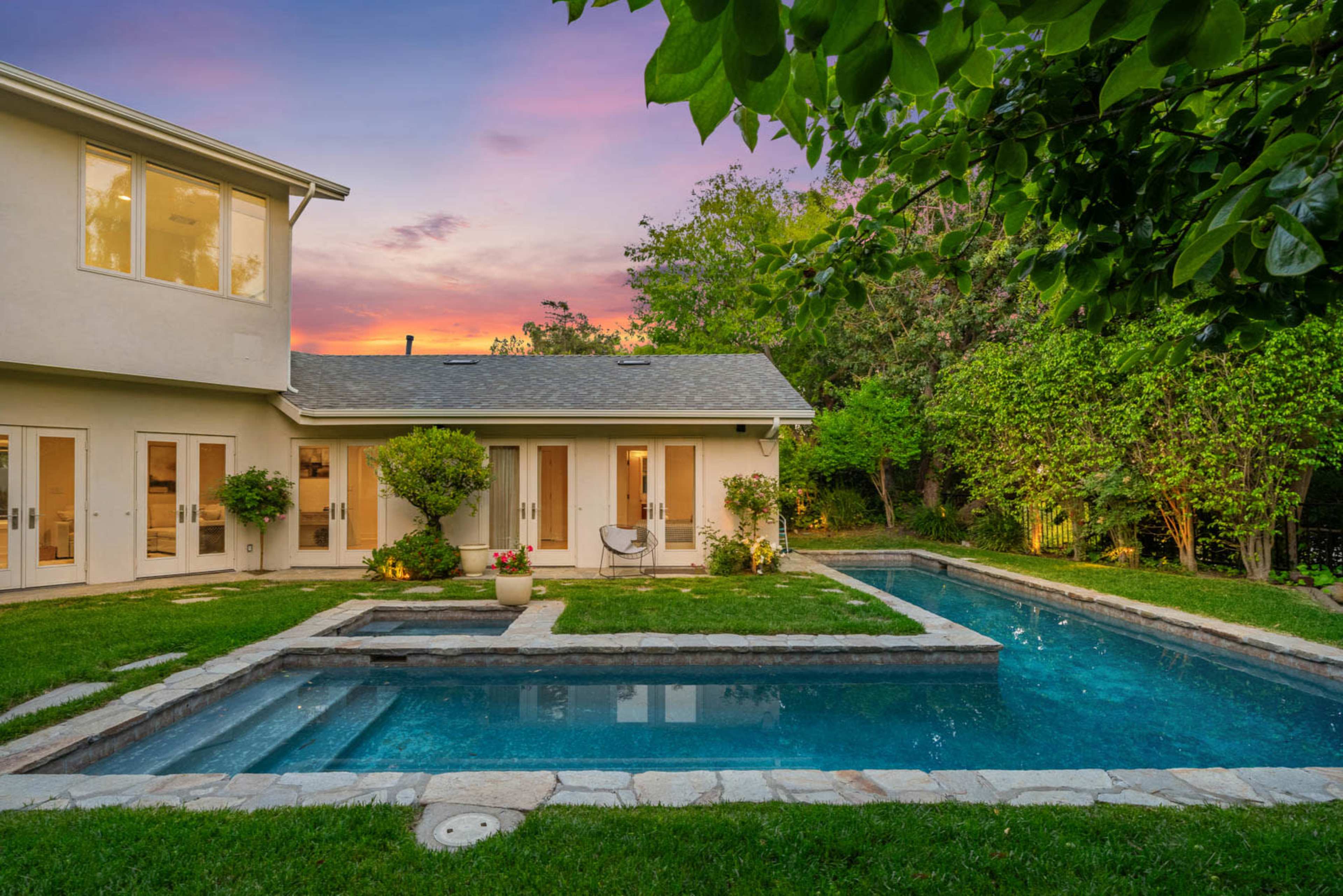 A well-maintained backyard features a swimming pool and a spa surrounded by green grass and trees under a colorful sky at sunset.