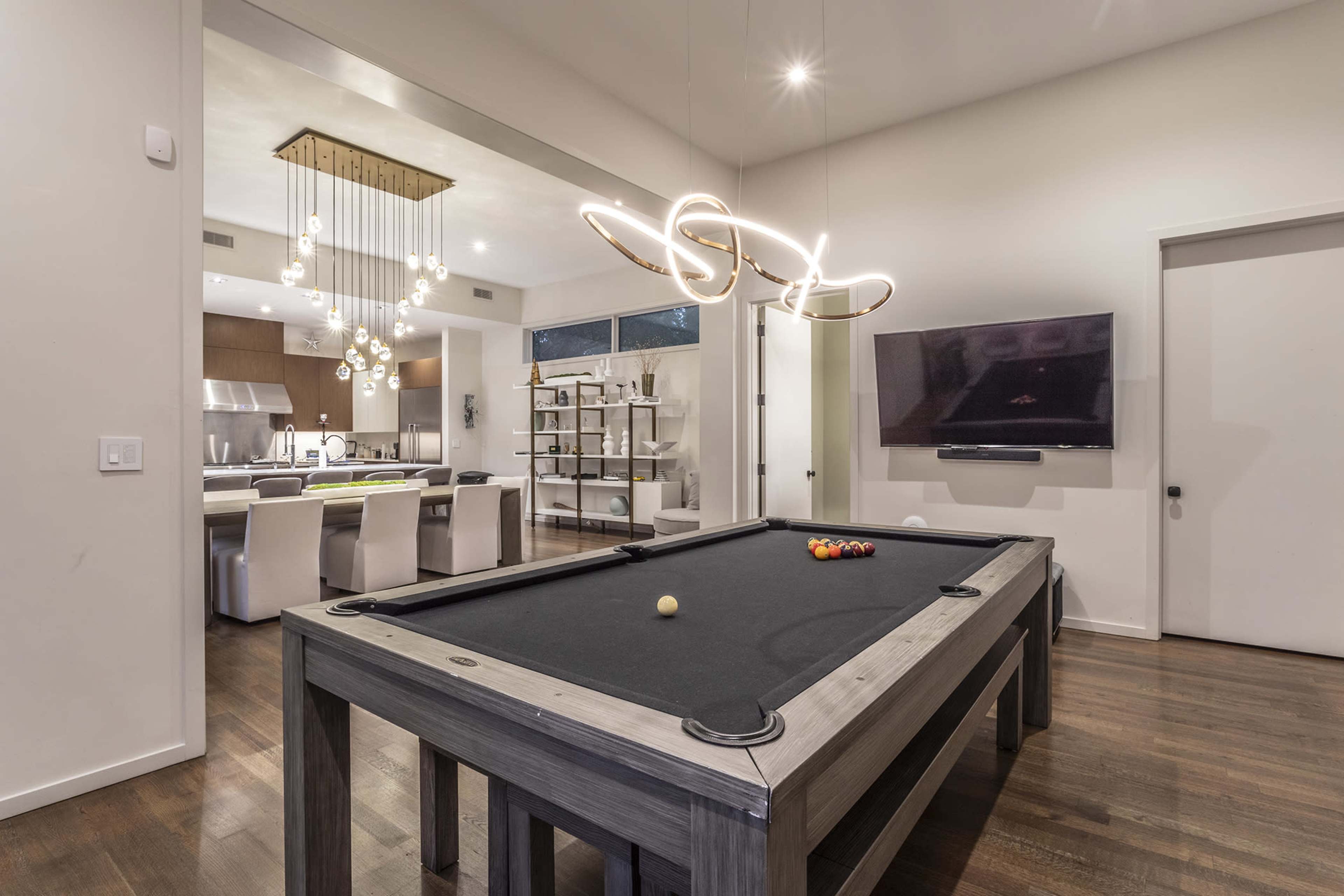 The image shows a modern living space featuring a pool table in the foreground, with an open kitchen and dining area visible in the background.