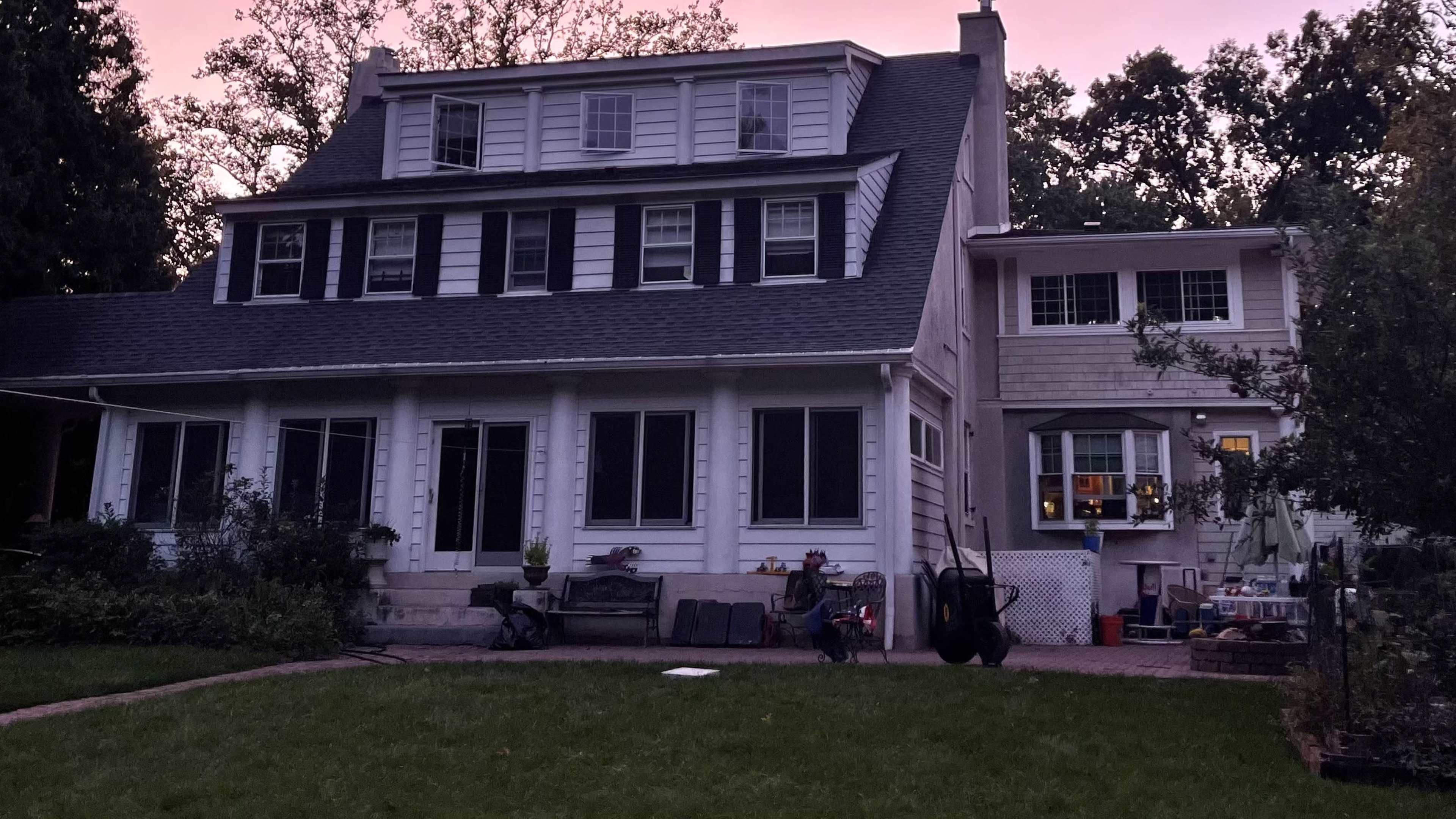 A two-story house with multiple windows and a porch is set against a twilight sky, surrounded by trees and a garden area.