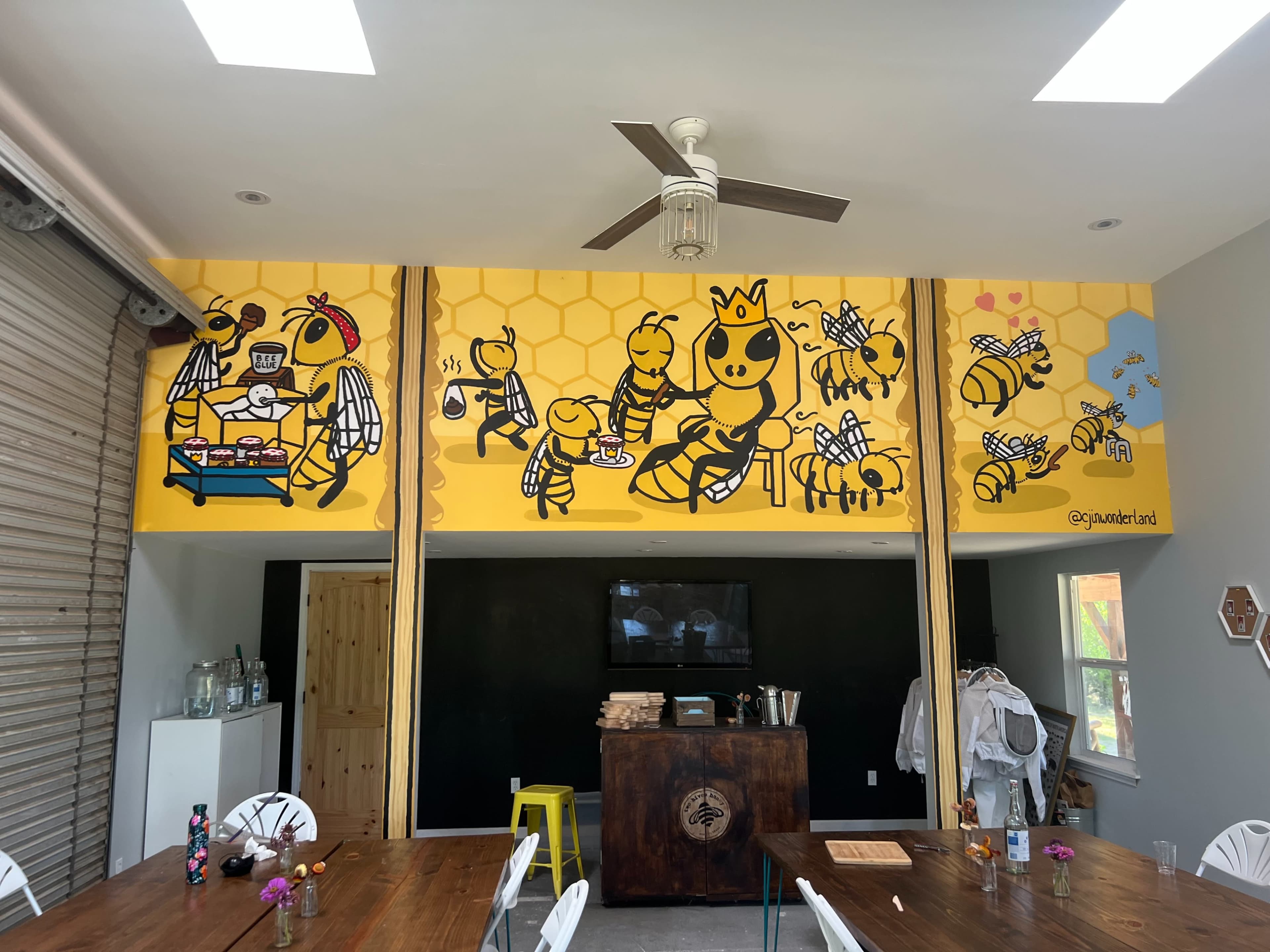 A bright mural of cartoon bees engaged in various activities on a yellow wall inside a café or workshop space.