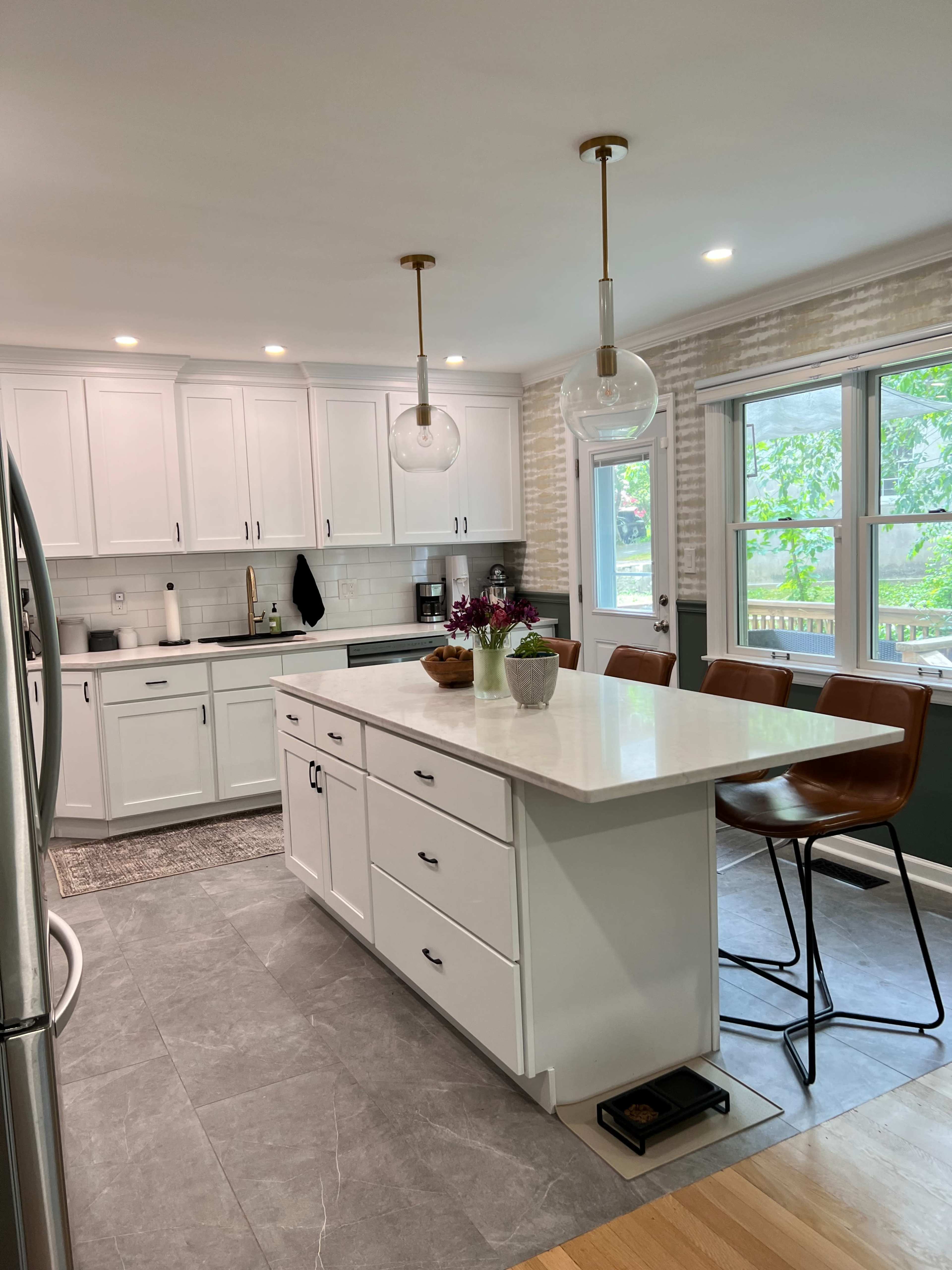 The image shows a modern kitchen featuring white cabinetry, a central island with seating, and large windows providing natural light.